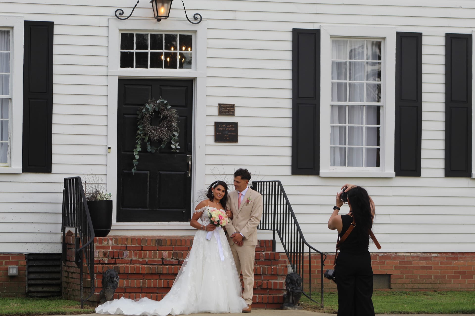 Bride in a white ball gown holding a pink bouquet and groom in a tan suit pose together on the brick front steps of the 1821 Scarborough House manor while a photographer captures their wedding portrait.