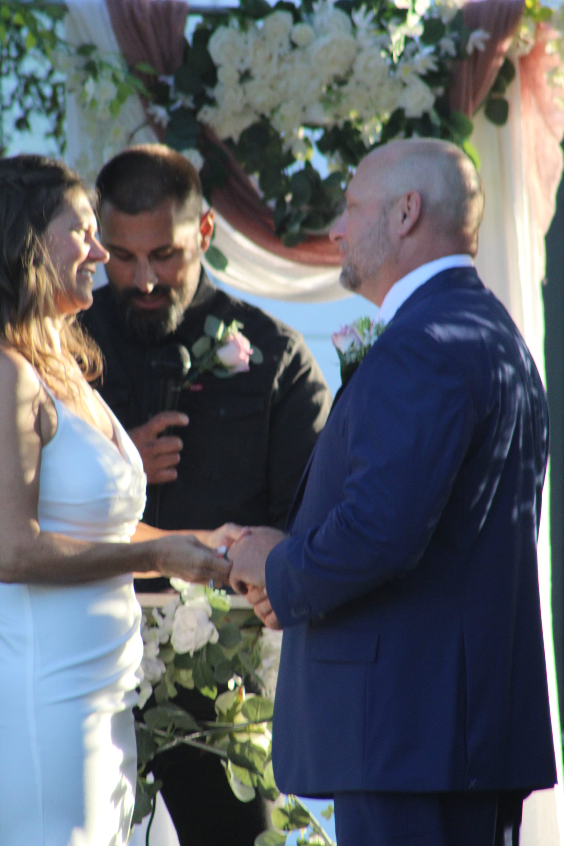 Bride in a white gown and groom in a navy suit exchange vows while holding hands beneath a lush white floral arch at Scarborough House.
