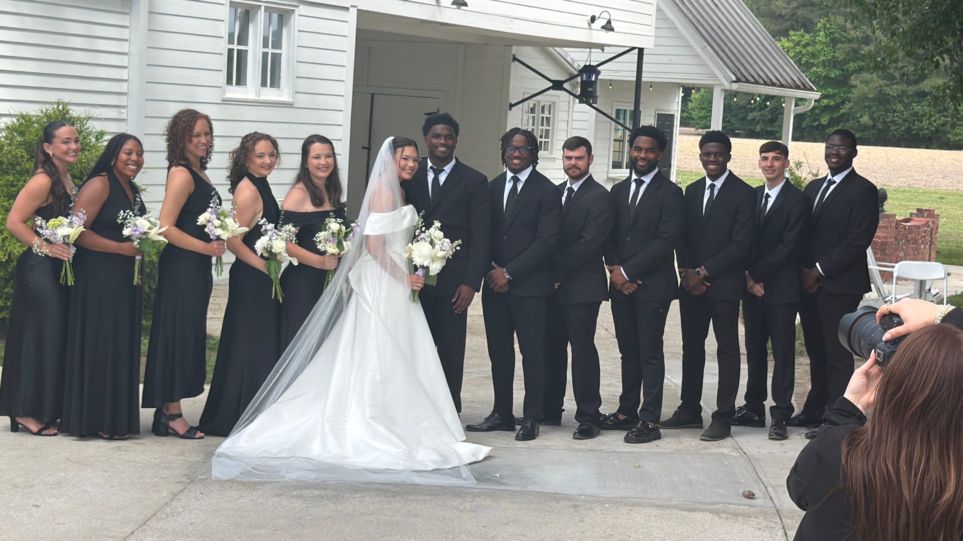 Bride in a white gown and veil poses with bridesmaids in black dresses and groomsmen in black suits in front of the historic white Scarborough House manor.