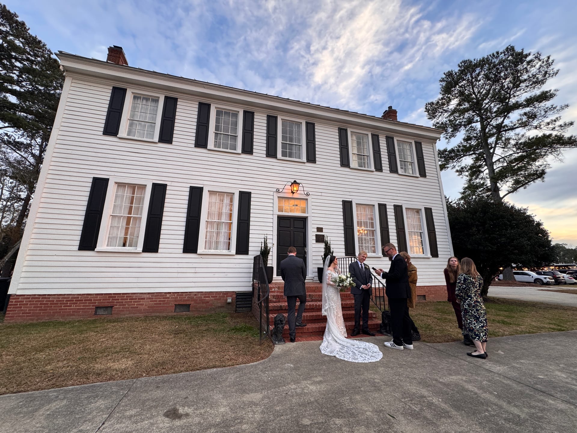 A bride in a lace gown with a cathedral train exchanges vows with her groom on the brick front steps of the 1821 Scarborough House manor at dusk, surrounded by a small group of witnesses.