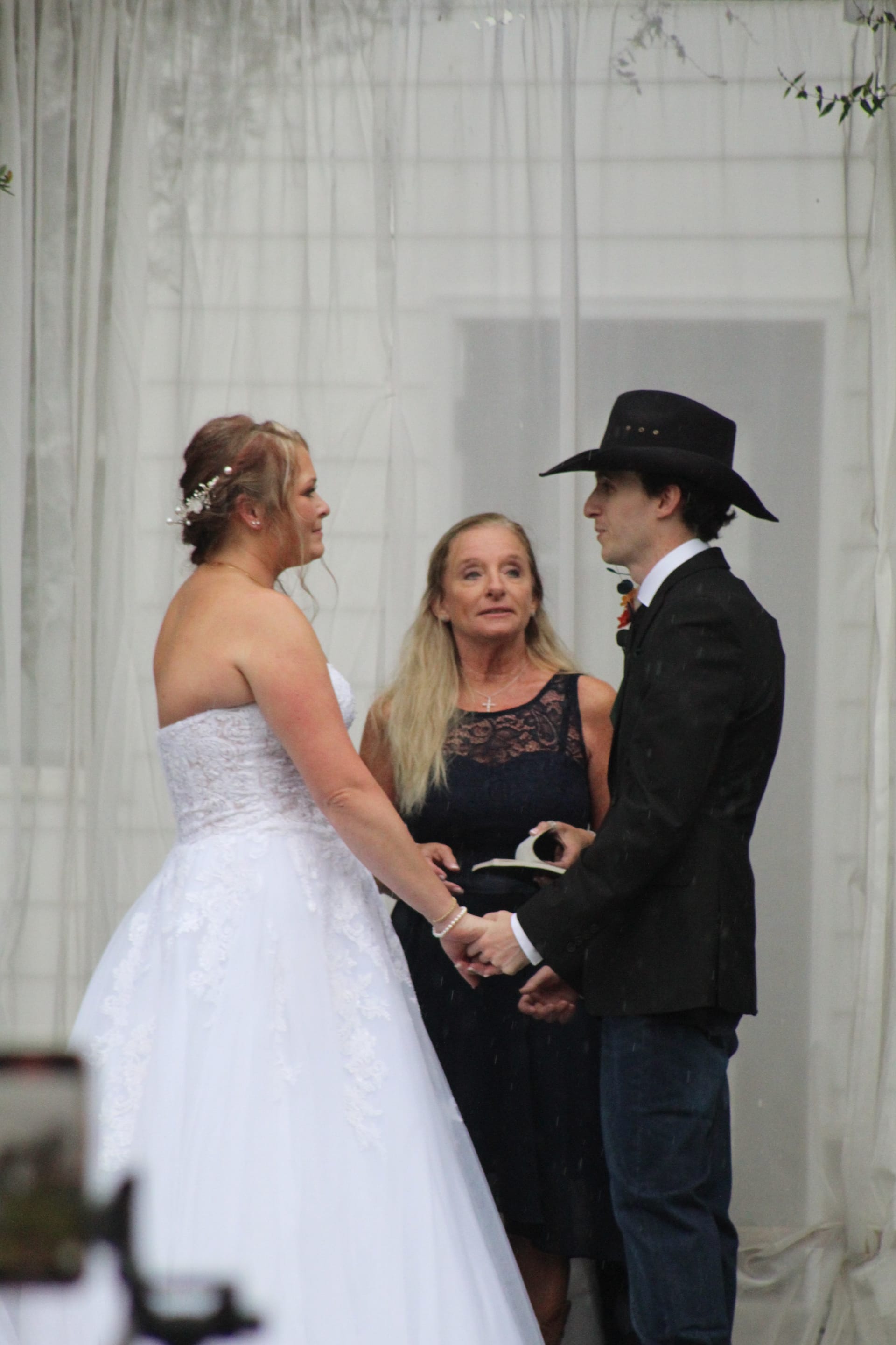 Bride in a strapless white lace gown and groom in a black suit and cowboy hat hold hands while exchanging vows with their officiant at Scarborough House wedding venue.