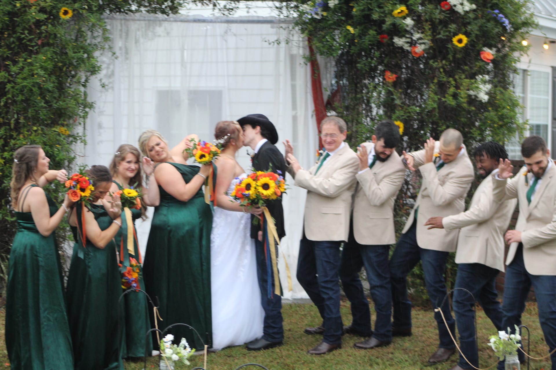 Bride and groom share their first kiss beneath a sunflower-adorned arch as bridesmaids in emerald gowns and groomsmen in cream blazers cheer on the ceremony lawn at Scarborough House.