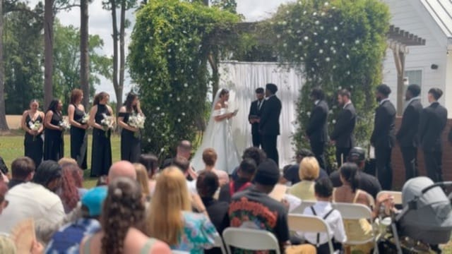 Outdoor wedding ceremony at Scarborough House with bride and groom at a lush greenery-covered arch surrounded by bridesmaids in black dresses, groomsmen, and seated guests on the ceremony lawn.