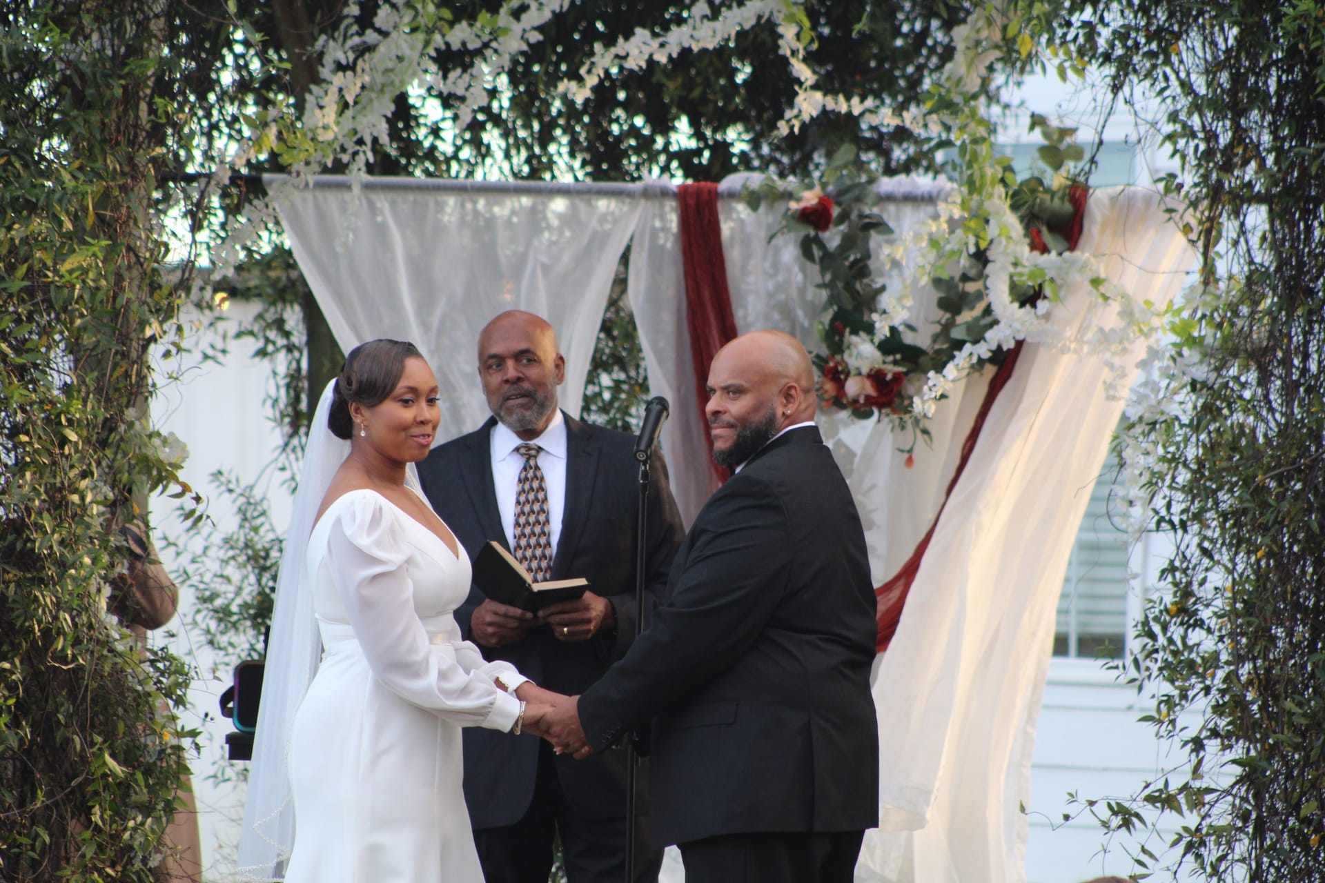 Bride in a white long-sleeve gown and groom in a black suit exchange vows while holding hands beneath a draped floral arch adorned with white flowers and burgundy accents at a garden ceremony.
