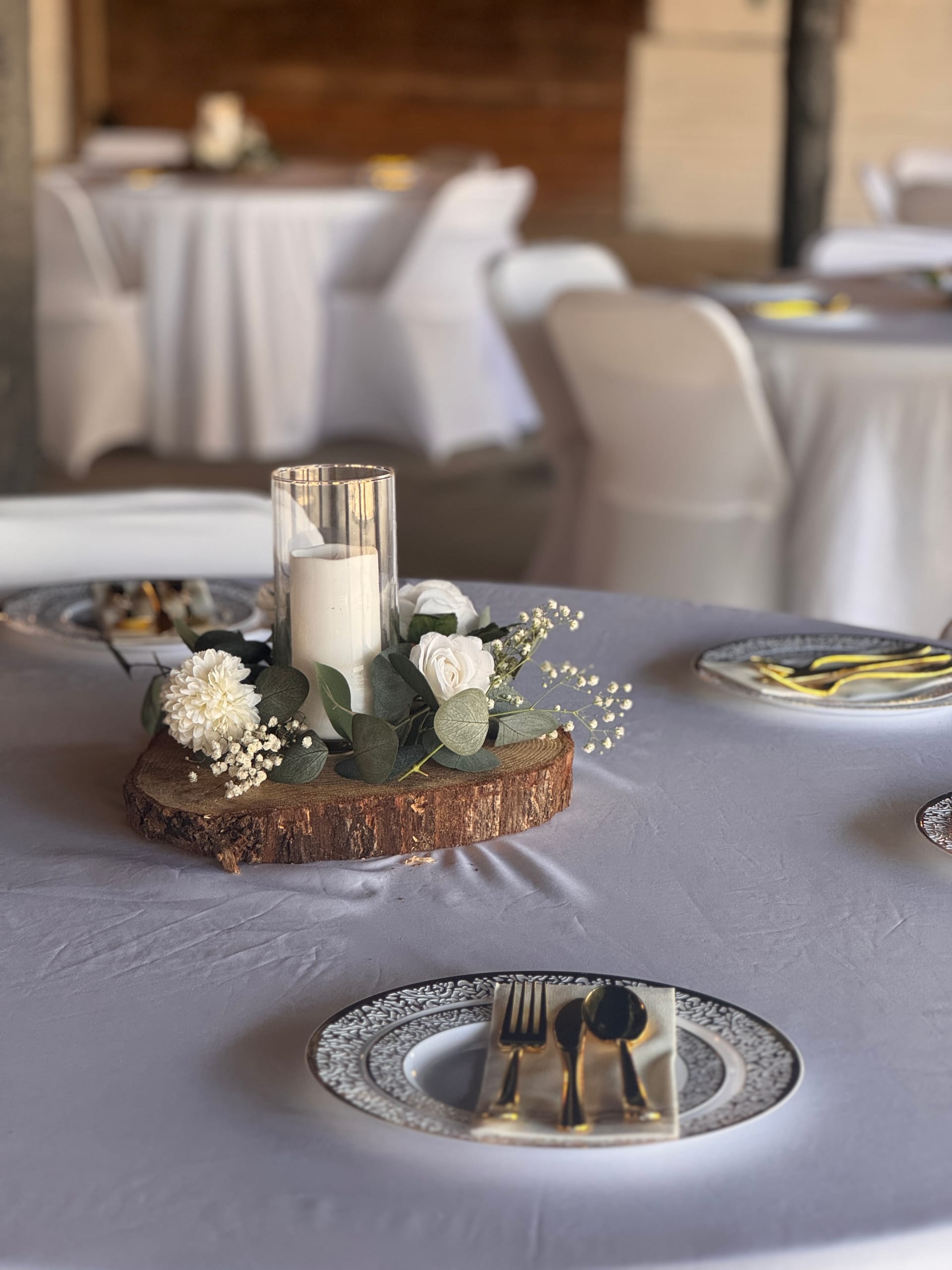 Elegant rustic wedding table centerpiece featuring a pillar candle in a glass hurricane vase surrounded by white roses, baby's breath, and eucalyptus on a natural wood slice, set on a gray linen tablecloth with patterned charger plates and gold cutlery.
