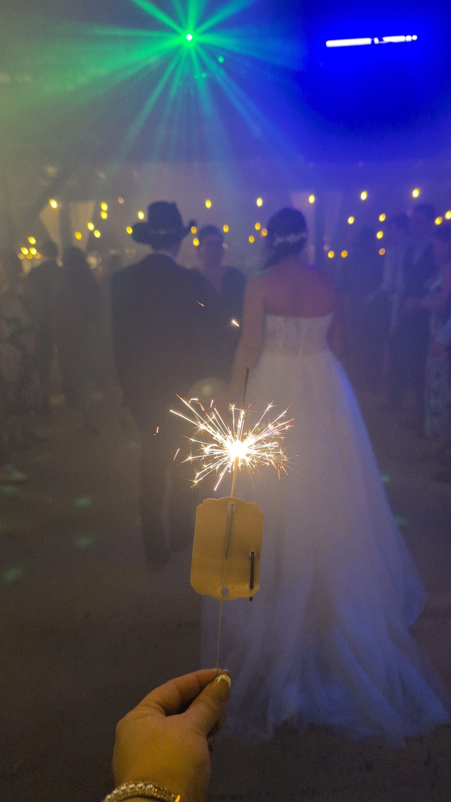 A glowing sparkler held in the foreground frames the silhouettes of a bride in a white gown and groom walking through a fog-filled reception hall bathed in colorful party lights at Scarborough House.