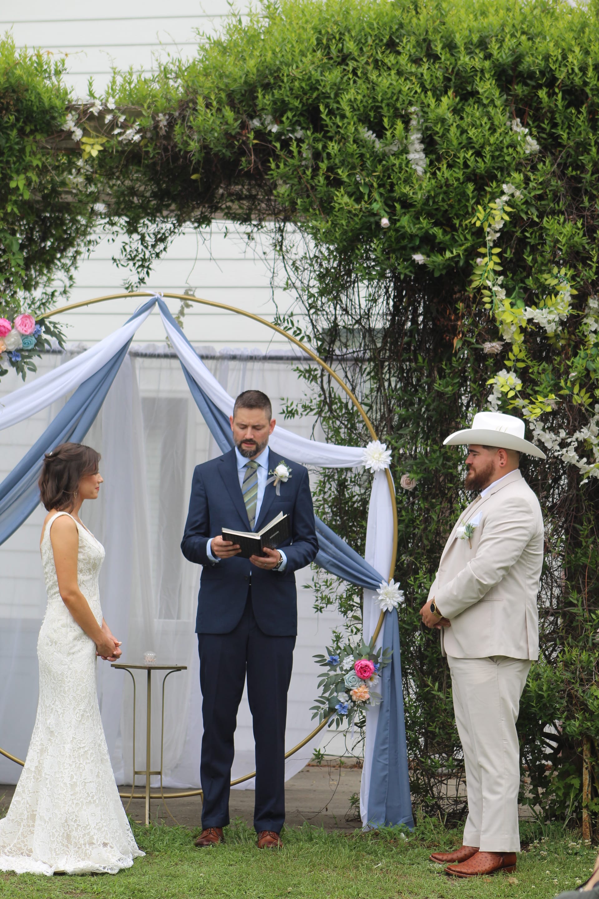 Bride in a lace gown and groom in a tan suit with white cowboy hat exchange vows beneath a gold hoop arch draped in dusty blue fabric and florals on the ceremony lawn at Scarborough House.