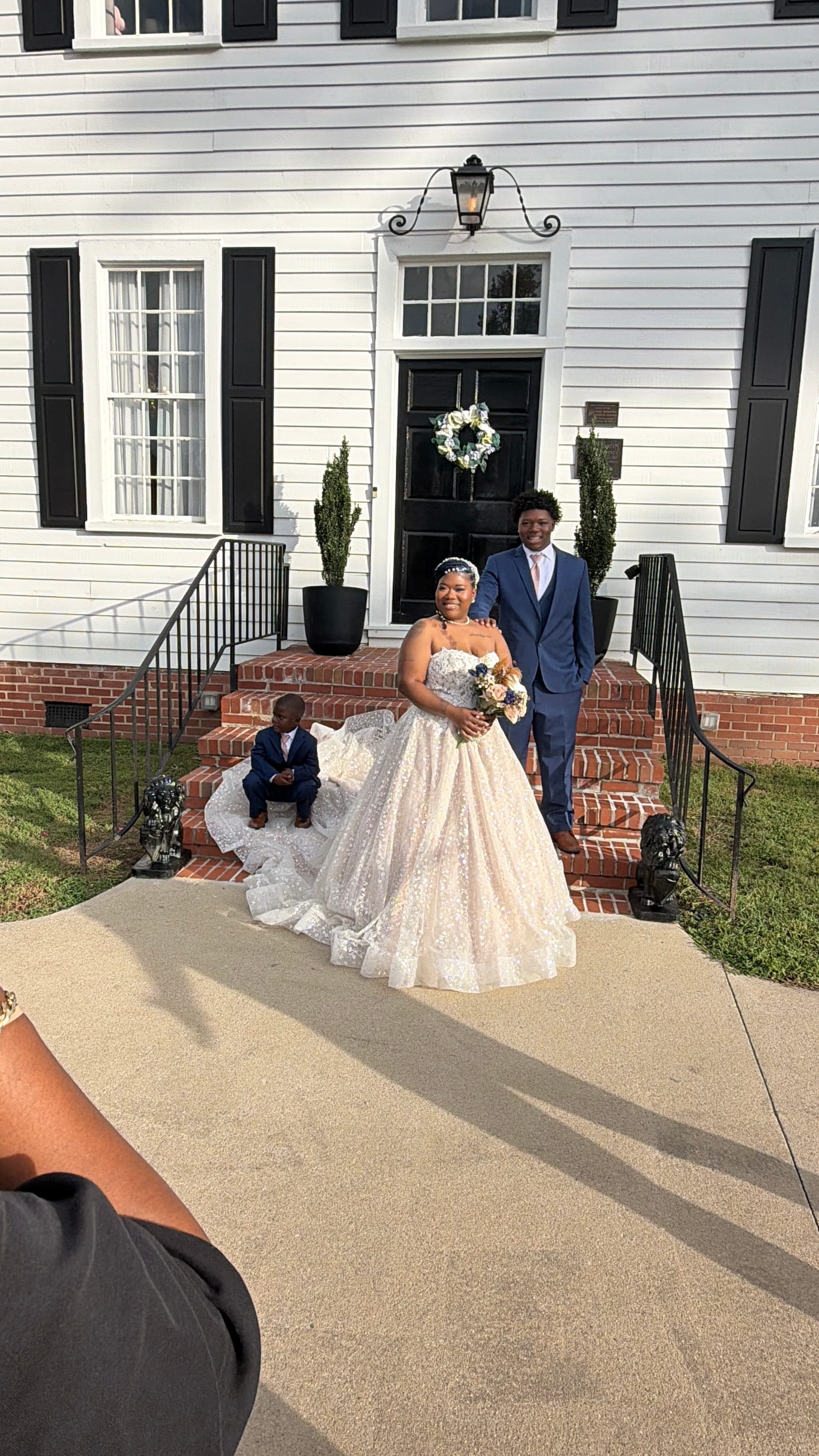 Bride in a full ballgown holding a bouquet and groom in a navy suit pose on the brick front steps of the historic 1821 Scarborough House manor while a young child in a matching suit sits on the steps behind them.
