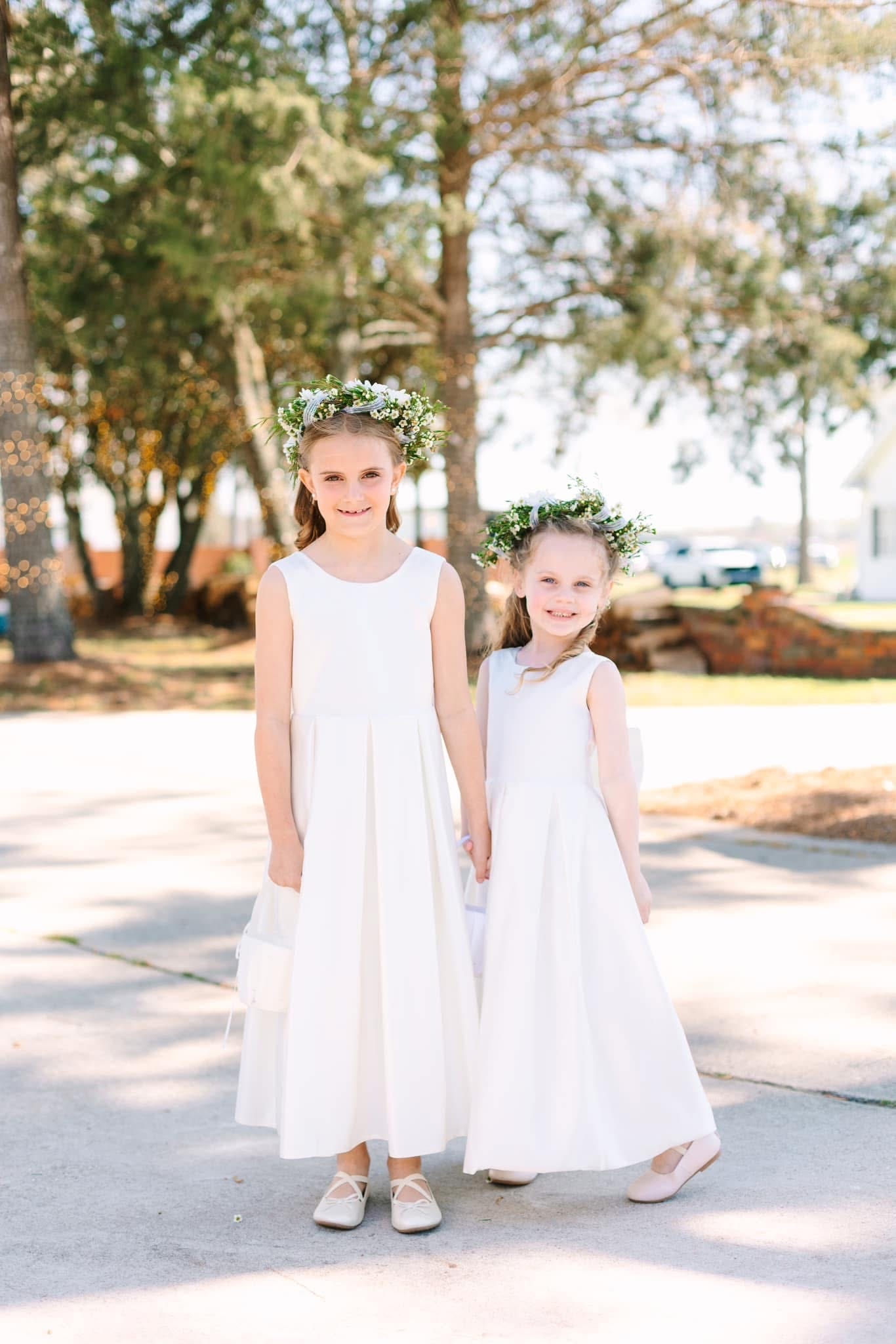 Two flower girls in long white dresses and greenery floral crowns hold hands and smile on the sun-dappled grounds of Scarborough House historic wedding venue.