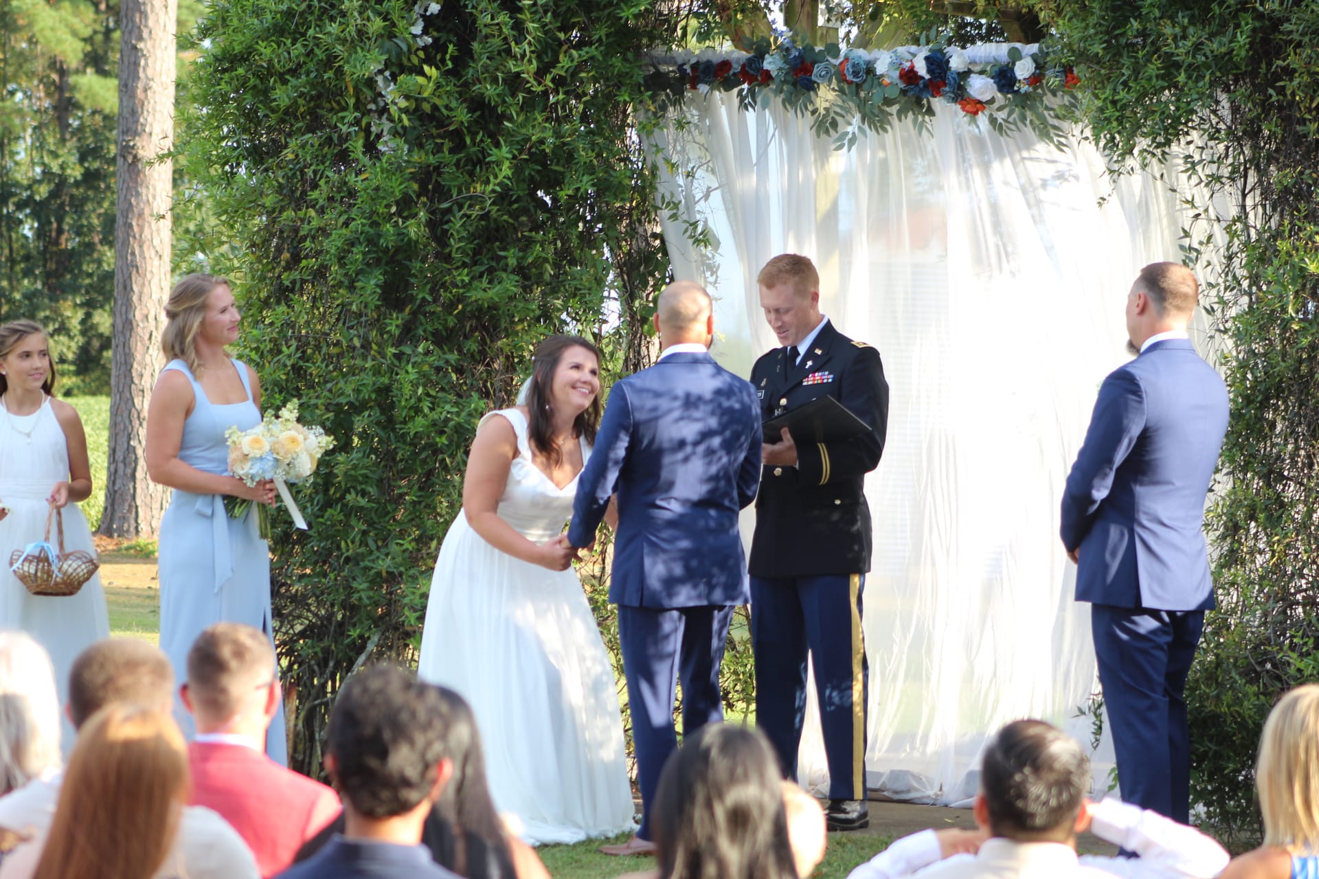 Smiling bride in a white gown holds hands with her groom beneath a lush green arch adorned with blue, red, and white flowers during an outdoor wedding ceremony at Scarborough House, with military and civilian groomsmen standing alongside.
