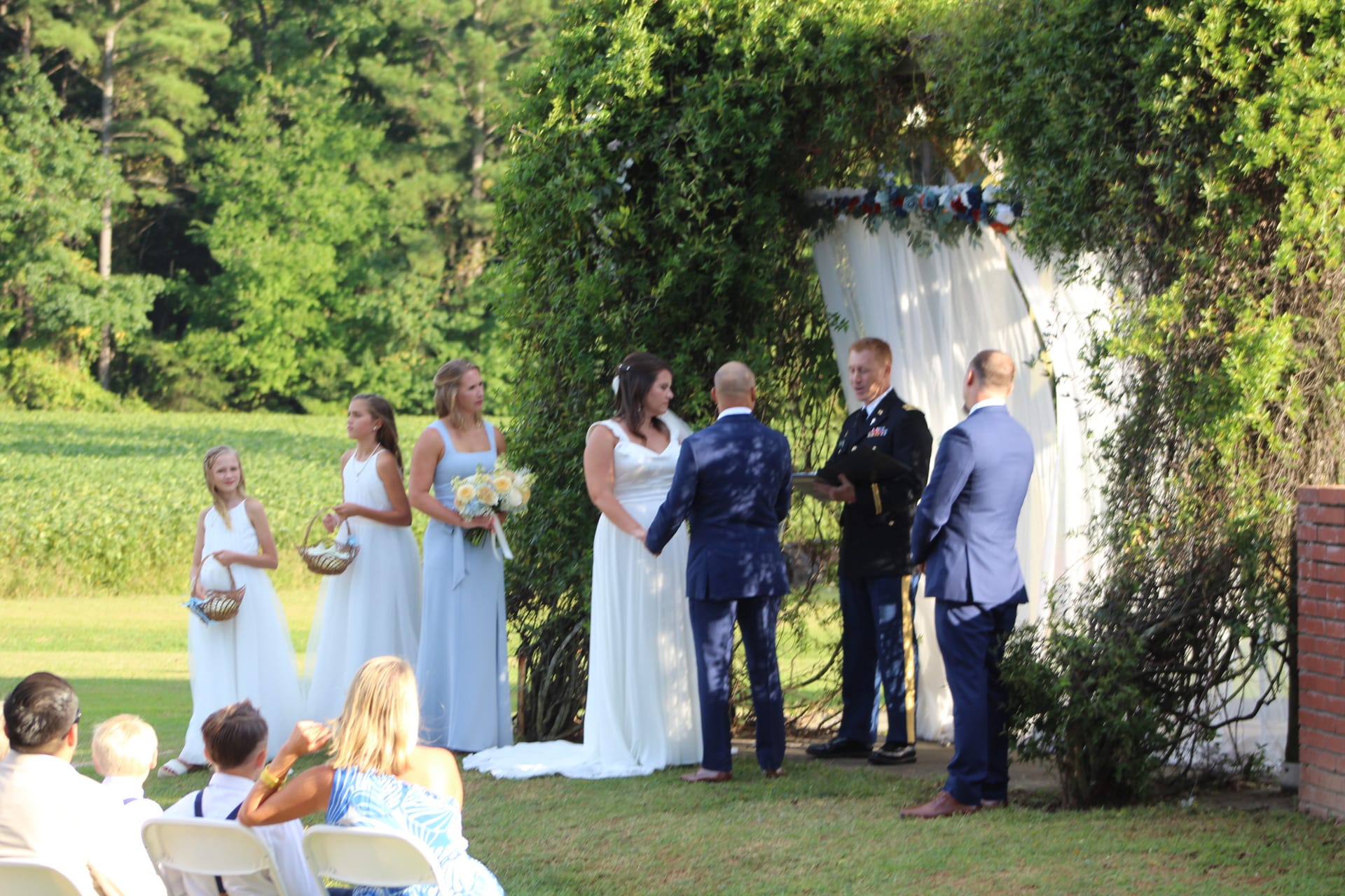 Bride and groom exchange vows beneath a lush vine-covered arch on the ceremony lawn at Scarborough House, flanked by bridesmaids in dusty blue gowns and a military officiant.