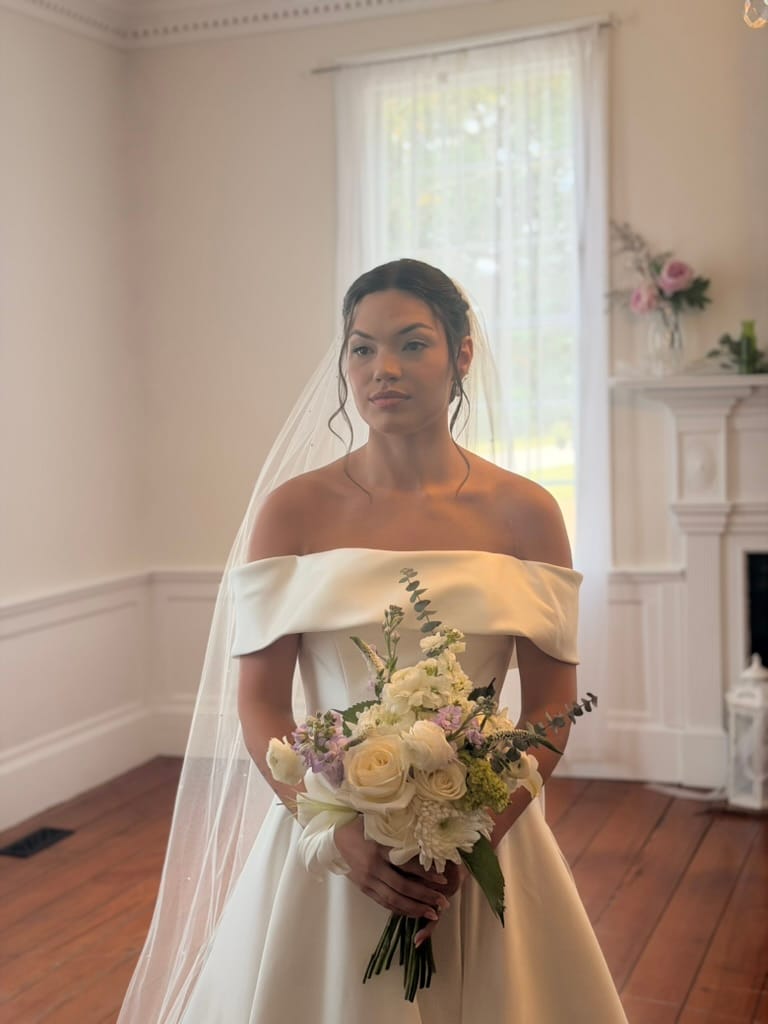 Bride in an off-shoulder white satin gown and cathedral veil holds a soft bouquet of cream roses and lavender blooms inside a light-filled historic room at Scarborough House.