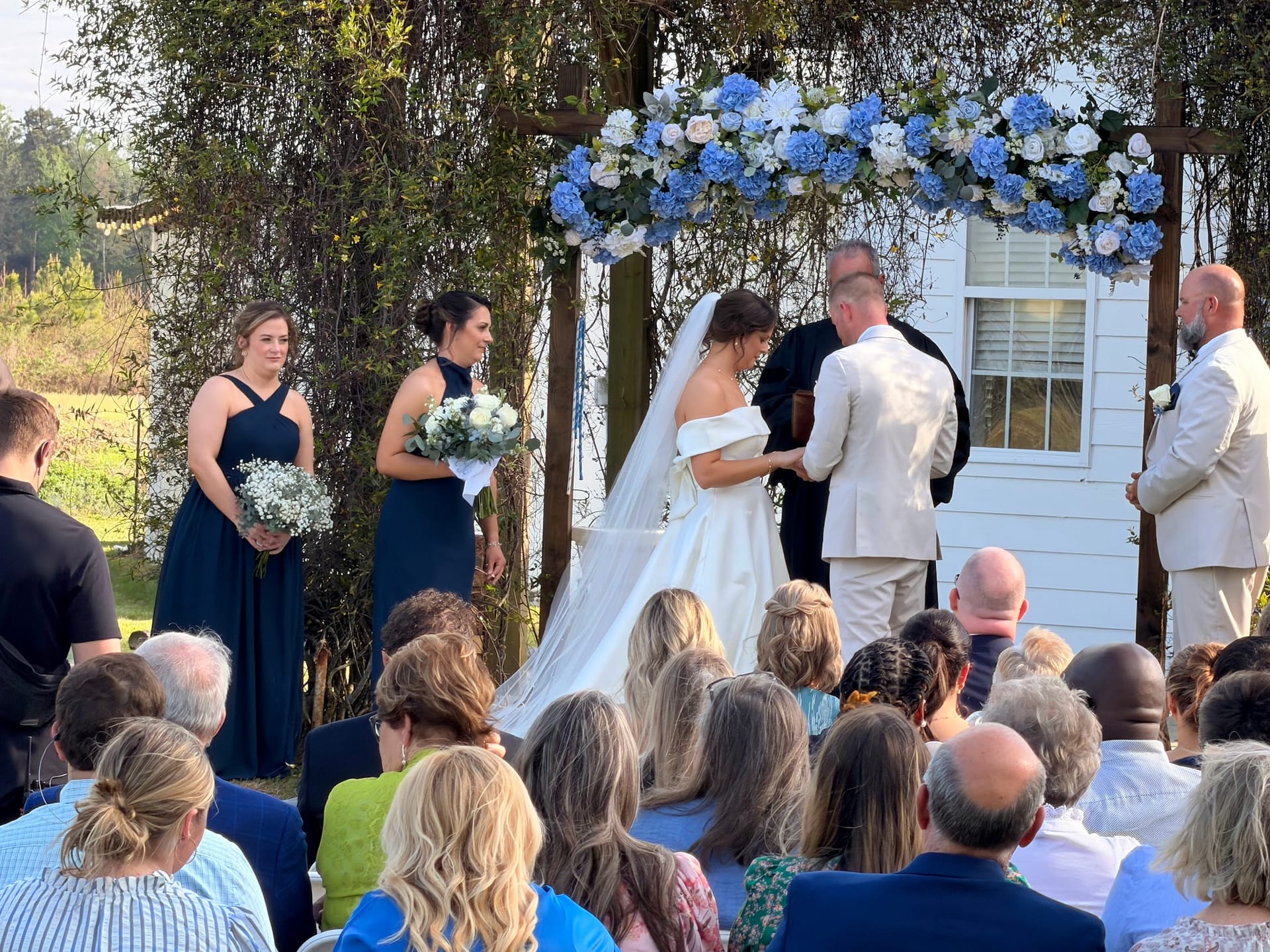 Bride and groom exchange vows beneath a stunning blue and white hydrangea floral arch on the ceremony lawn at Scarborough House, flanked by bridesmaids in navy and surrounded by seated guests.