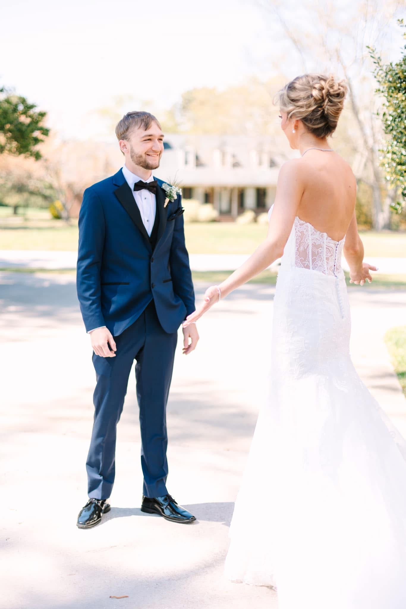 Groom in a navy tuxedo smiles joyfully as he sees his bride in a backless lace gown for the first time on the sunlit grounds of Scarborough House historic estate.