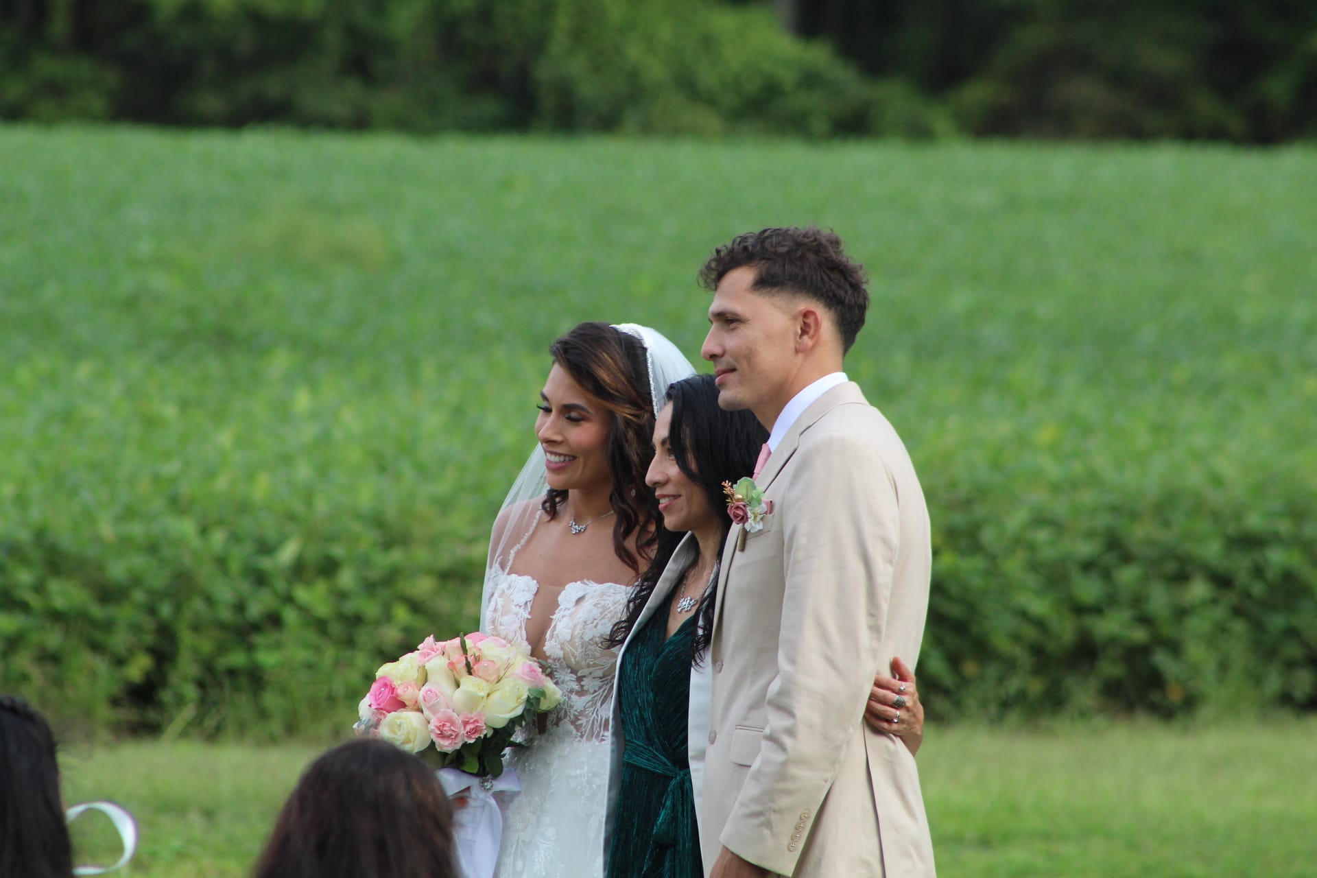 Smiling bride in lace gown holding pink and white bouquet stands beside a woman in a dark green dress and groom in a tan suit with boutonniere on the ceremony lawn at Scarborough House with lush green fields in the background.