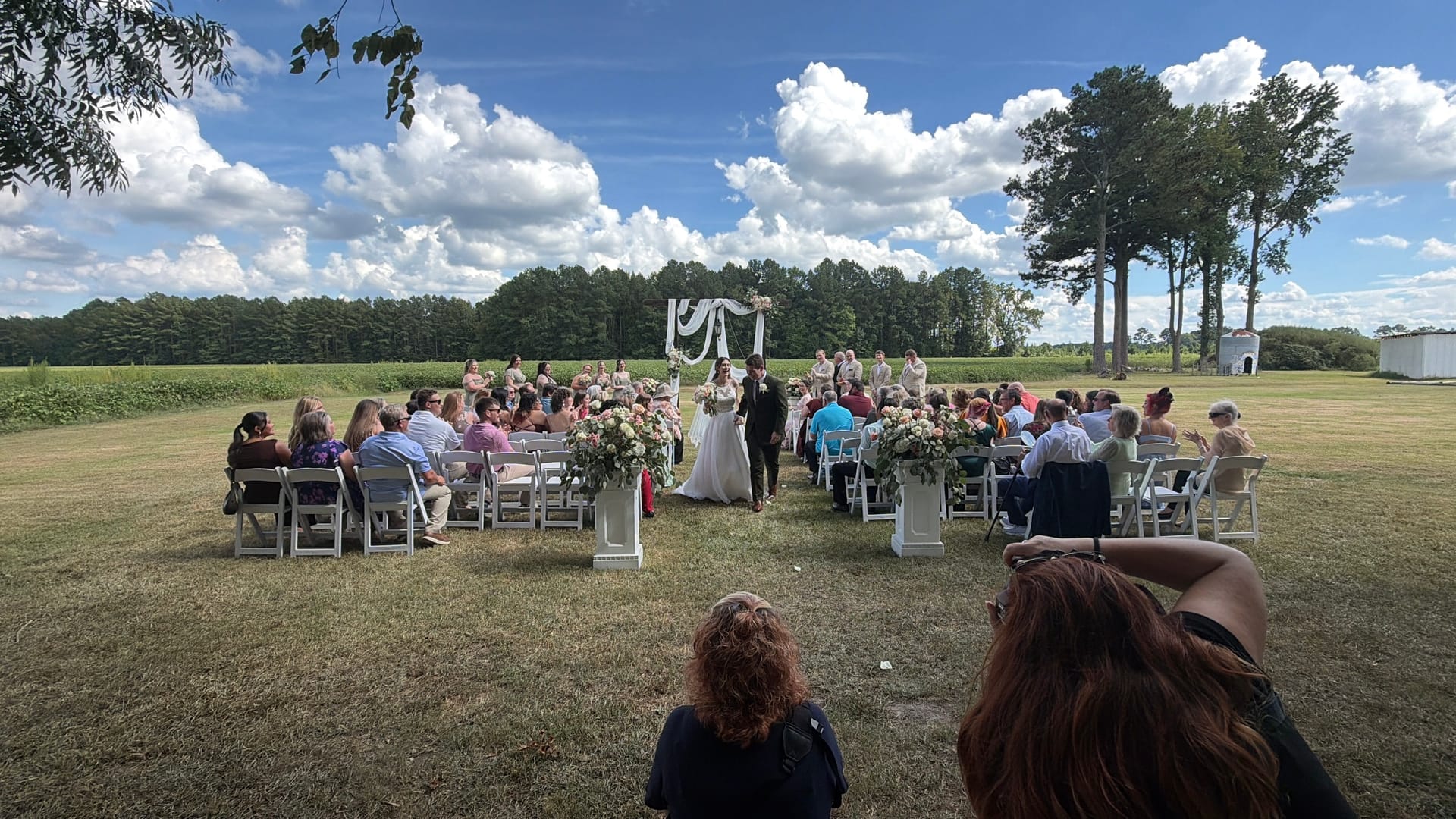 Outdoor wedding ceremony on the Scarborough House ceremony lawn with guests seated in white chairs flanking a floral-lined aisle leading to a draped white arch under a bright blue sky.