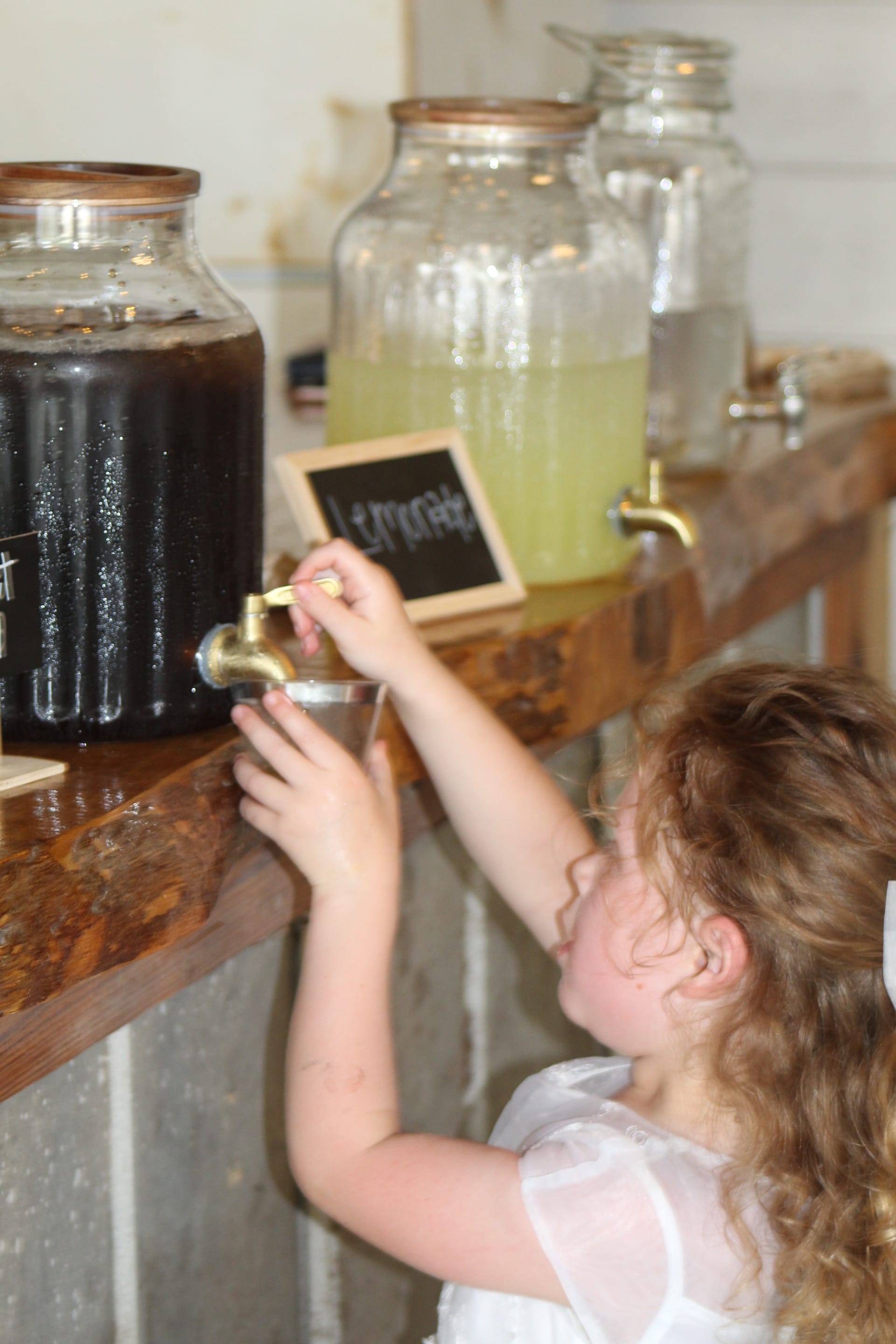 A young flower girl in a white dress reaches up on tiptoe to pour a drink from a brass-tapped glass dispenser at a rustic wooden beverage bar labeled Lemonade during a wedding reception.
