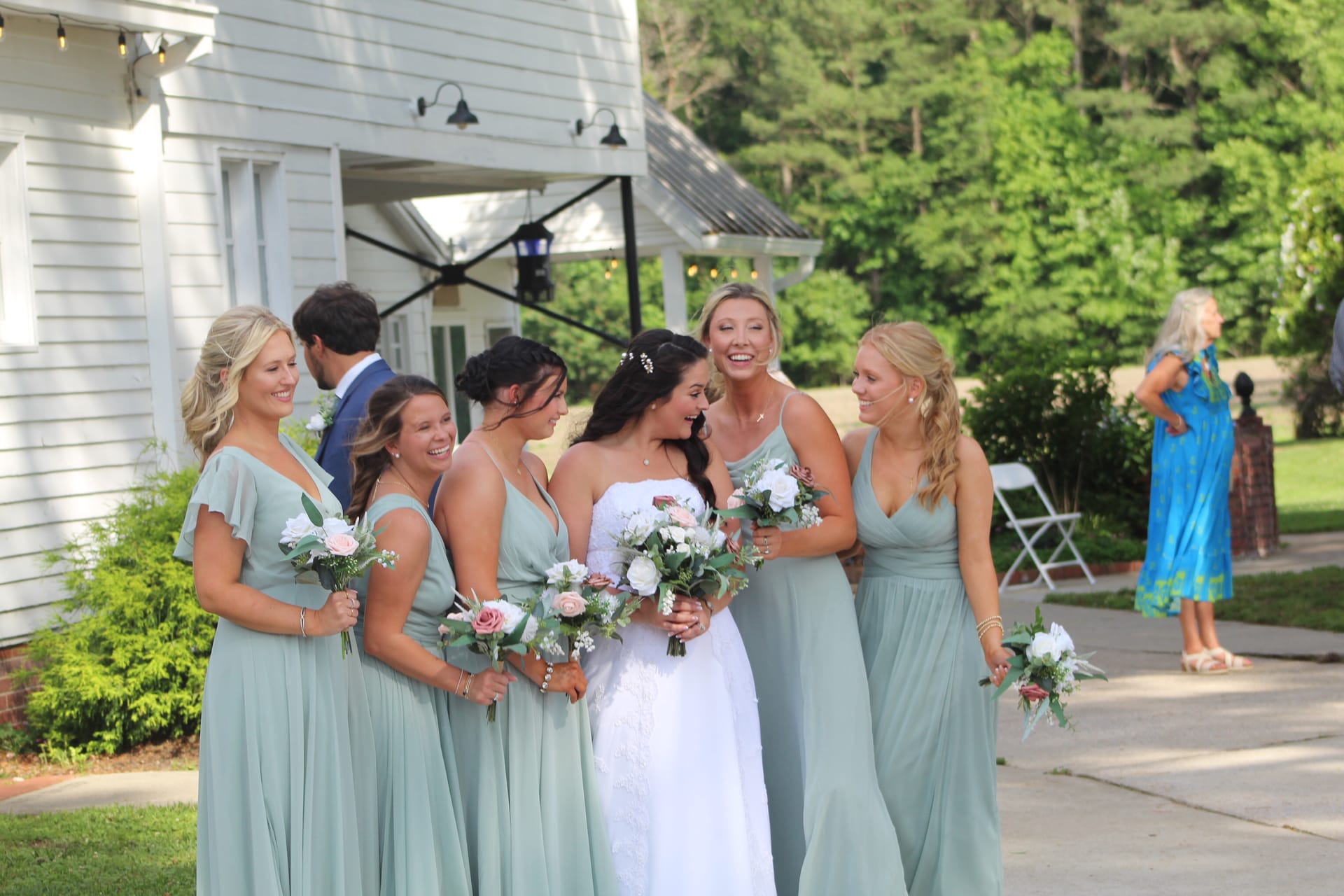 Bride in white lace gown laughing with five bridesmaids in sage green dresses holding pink and white bouquets outside a historic white manor house.
