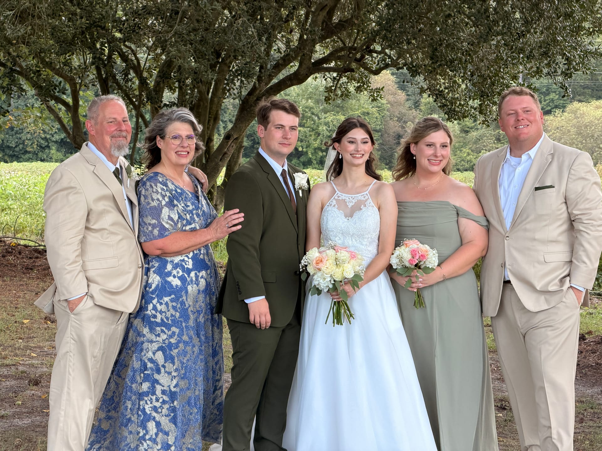 Bride in white gown holding a bouquet of roses poses with five family members including groom in olive suit under sweeping oak trees at Scarborough House estate wedding venue.