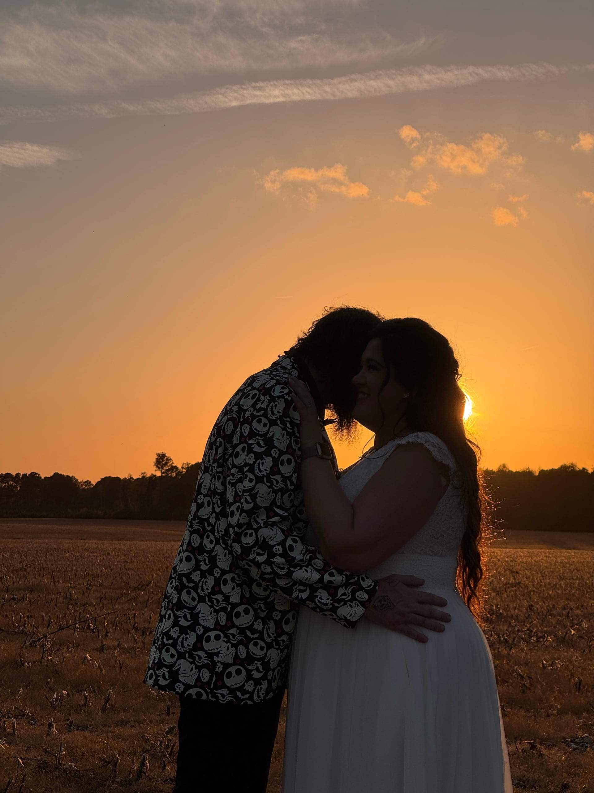 Bride in white gown and groom in Nightmare Before Christmas blazer share a kiss silhouetted against a vivid orange sunset over open farmland fields at Scarborough House.
