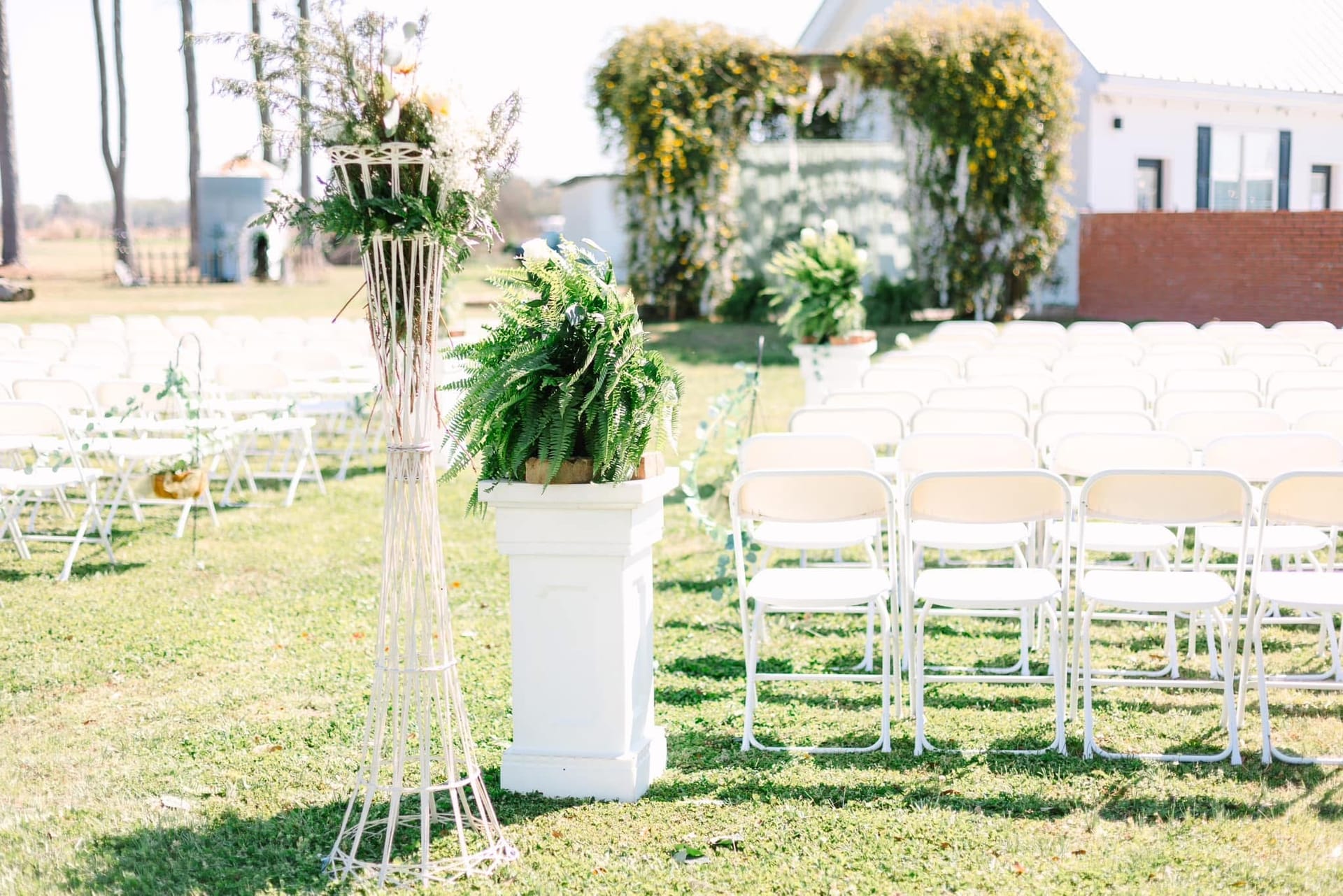 Outdoor wedding ceremony setup at Scarborough House featuring rows of white folding chairs, a potted Boston fern on a white pedestal, a decorative wicker plant stand, and a lush greenery-draped arch in the background.
