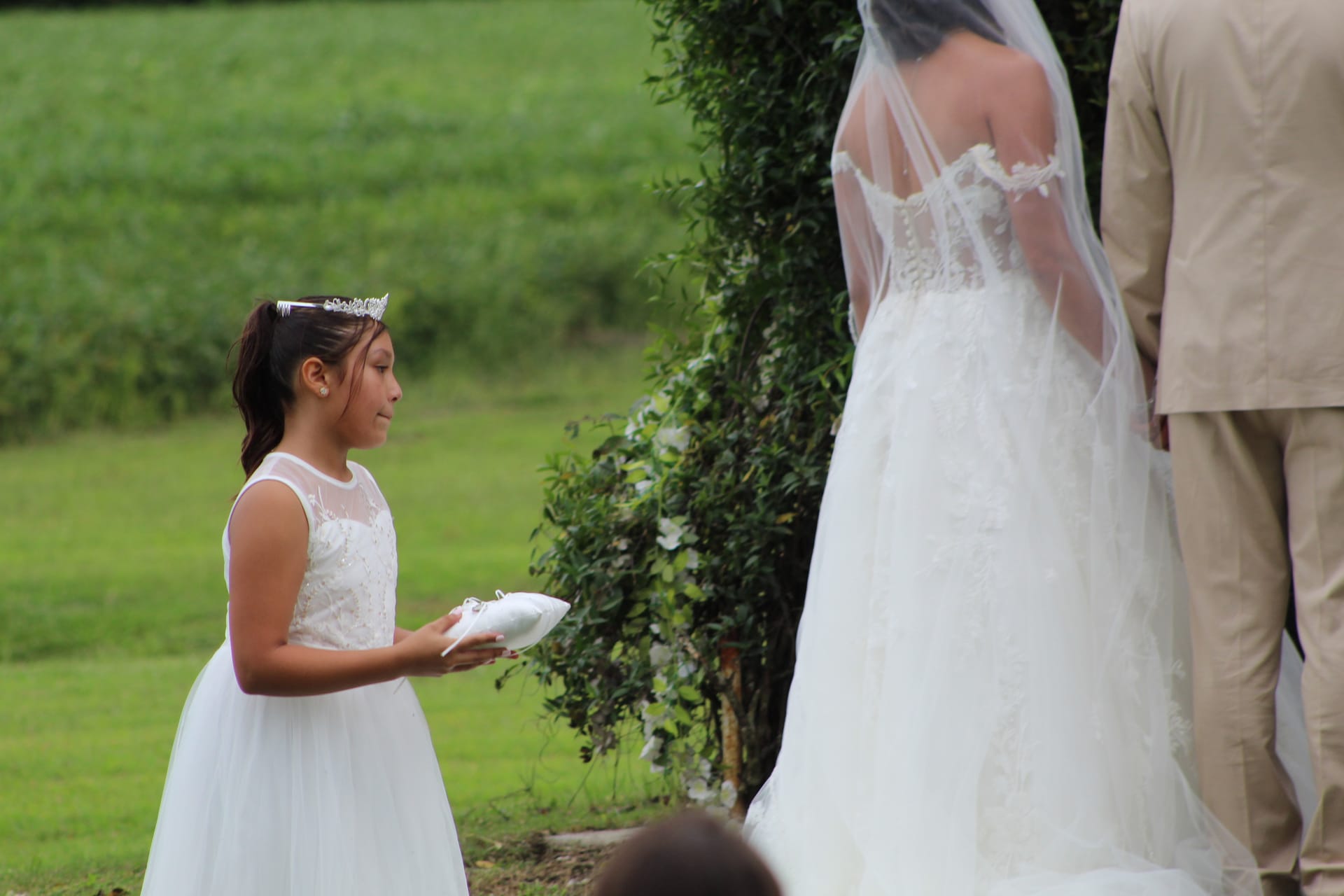 A young flower girl in a white dress and tiara presents the wedding ring pillow to the bride during an outdoor ceremony at Scarborough House, with lush greenery and farmland fields in the background.