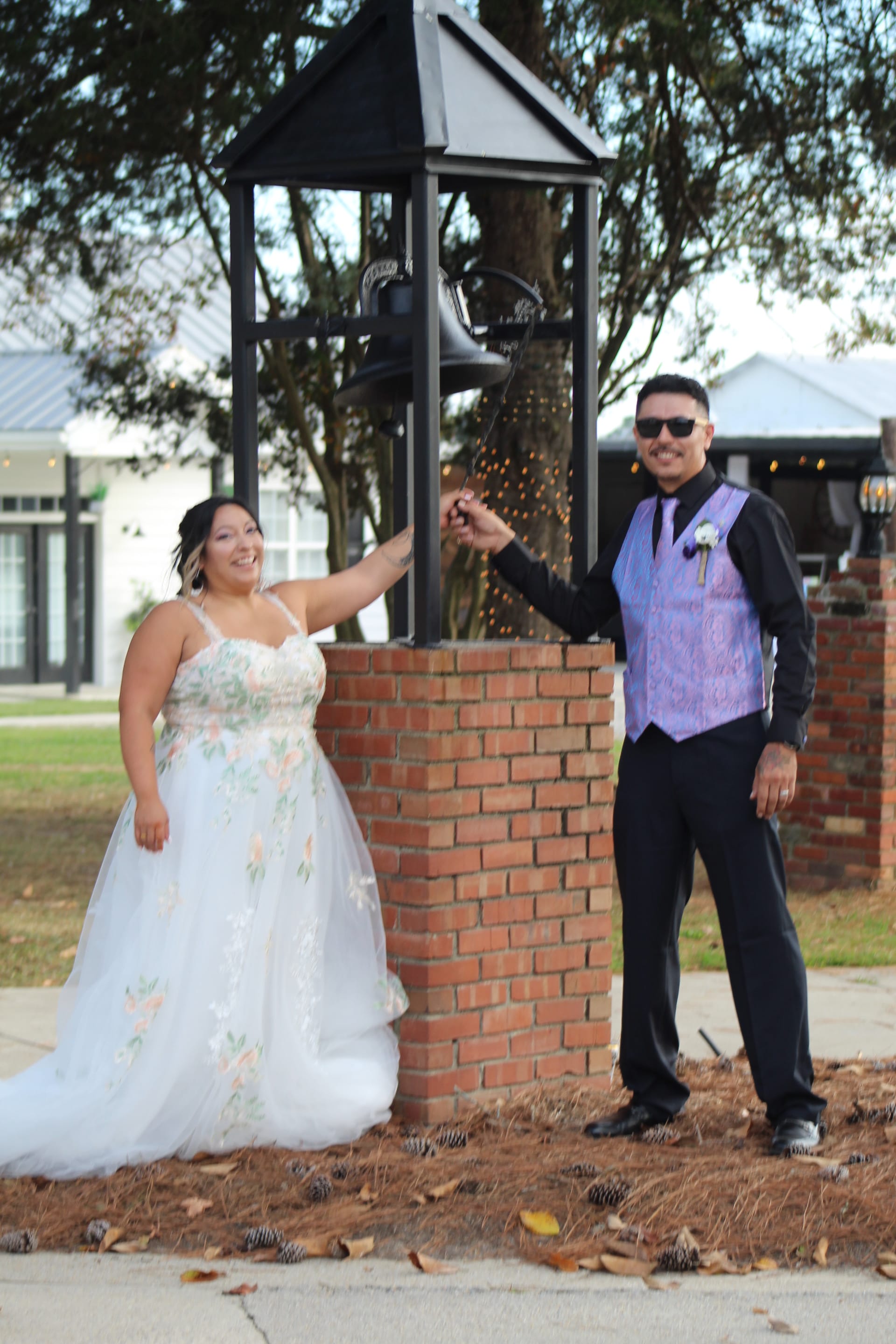 Bride in a floral ball gown and groom in a black shirt with purple vest ring the decorative bell together on the grounds of Scarborough House wedding venue.