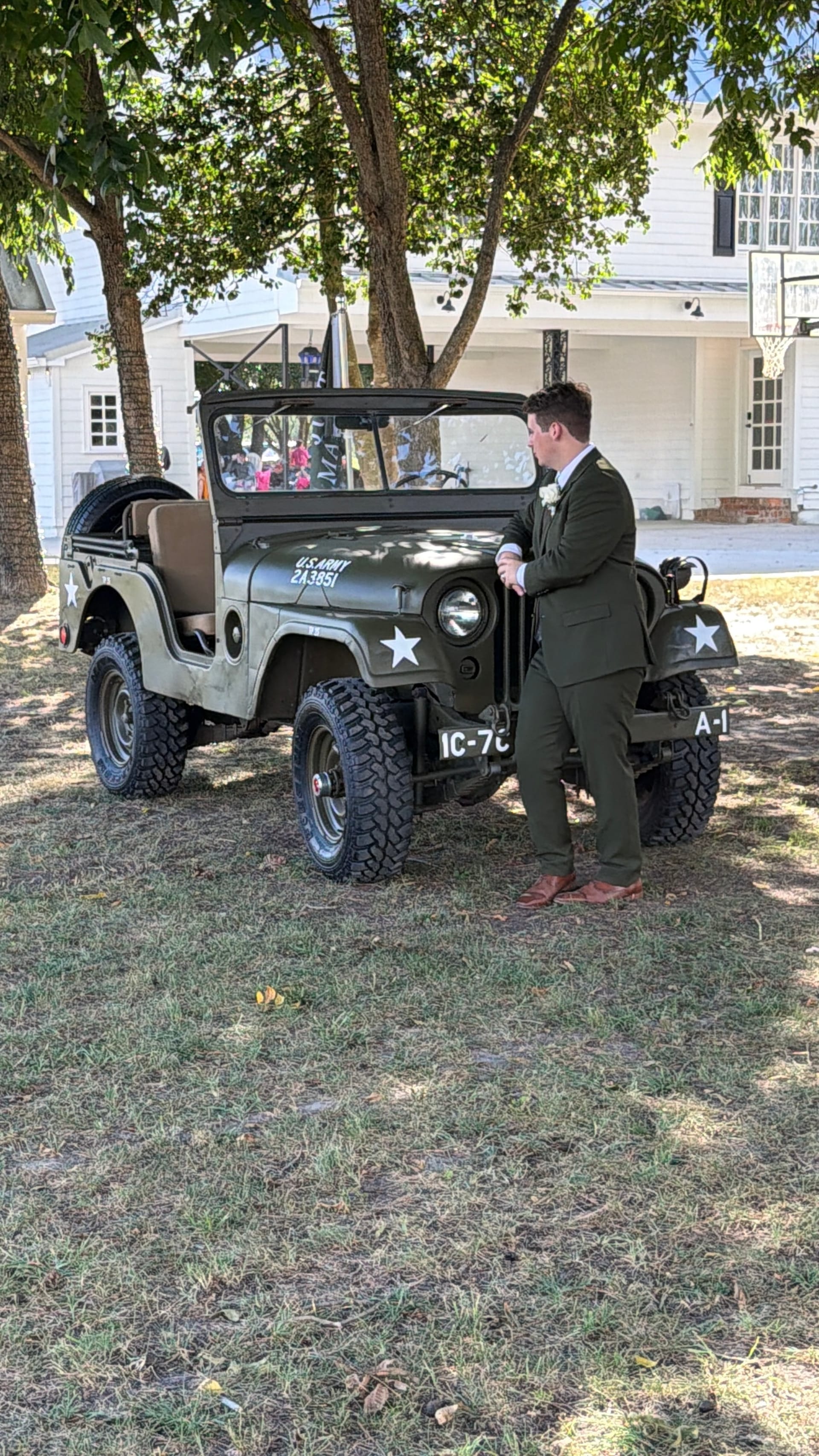 Groom in an olive green suit leans casually against a vintage U.S. Army Jeep beneath oak trees at Scarborough House historic estate wedding venue.