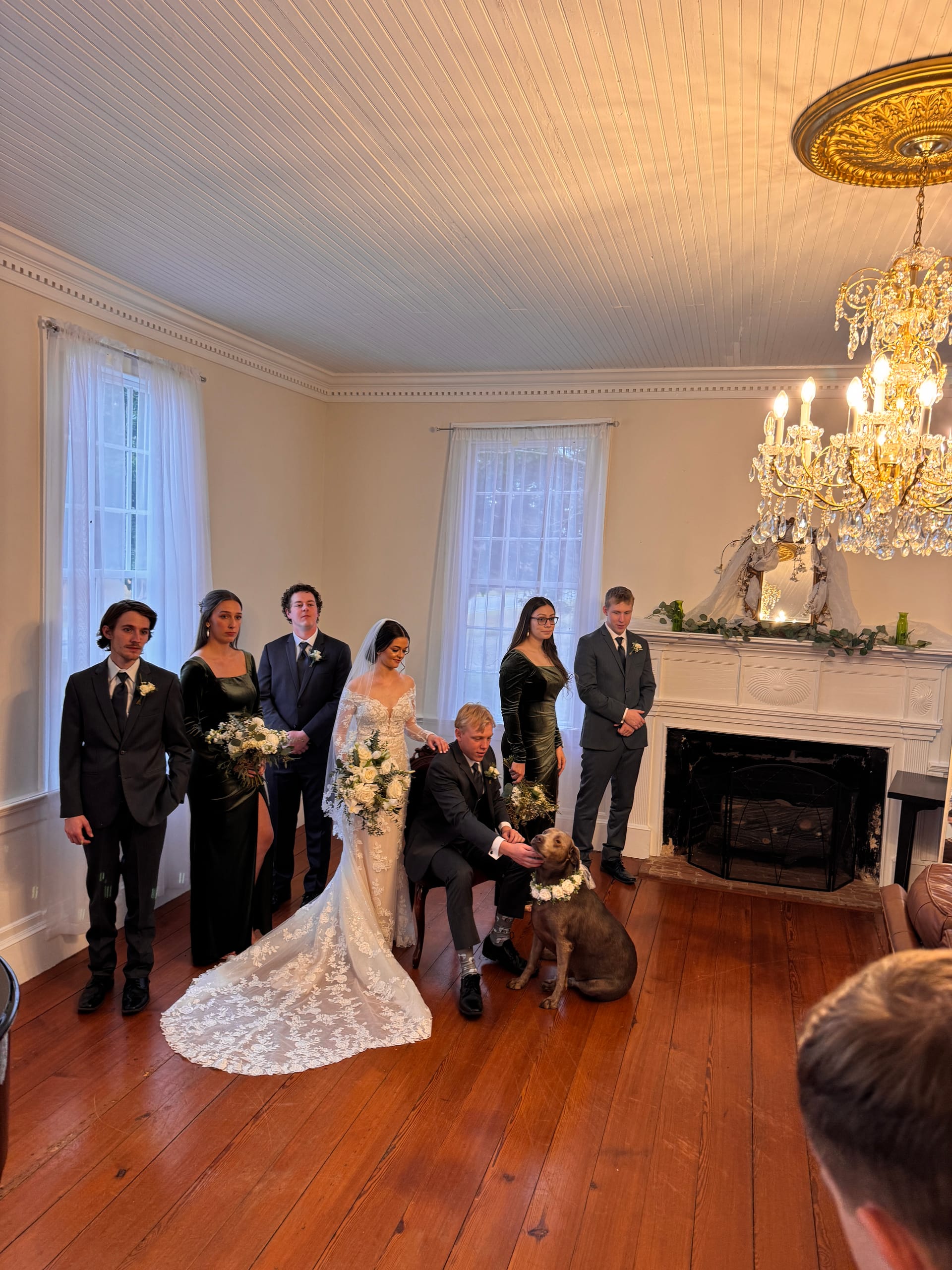 Bride in lace gown and groom crouching beside their flower-crowned dog surrounded by bridesmaids in black velvet and groomsmen in dark suits inside the chandelier-lit grand hall at Scarborough House.