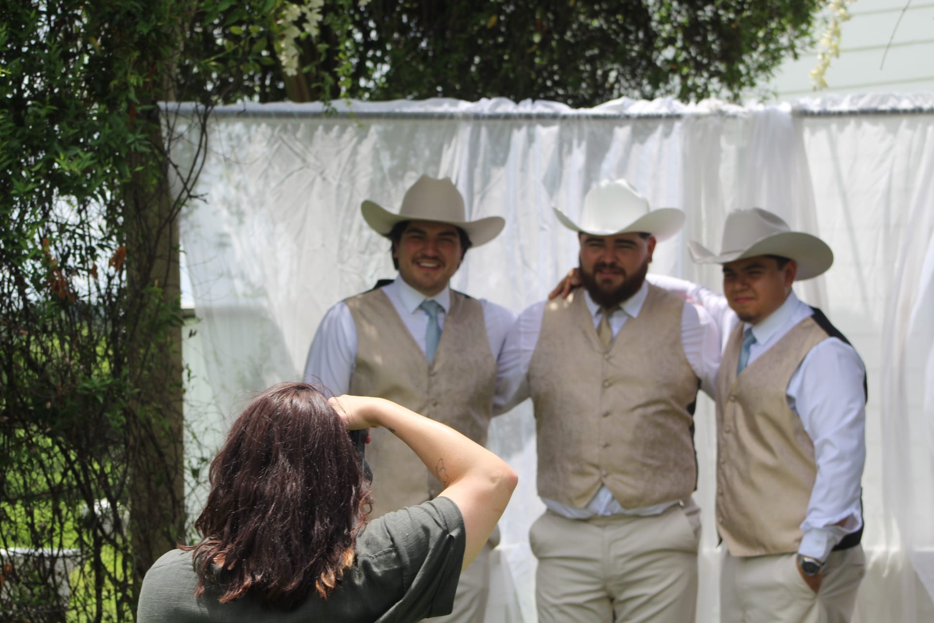 A photographer captures three smiling groomsmen wearing tan vests and cowboy hats posing together in front of a white draped backdrop at an outdoor wedding.