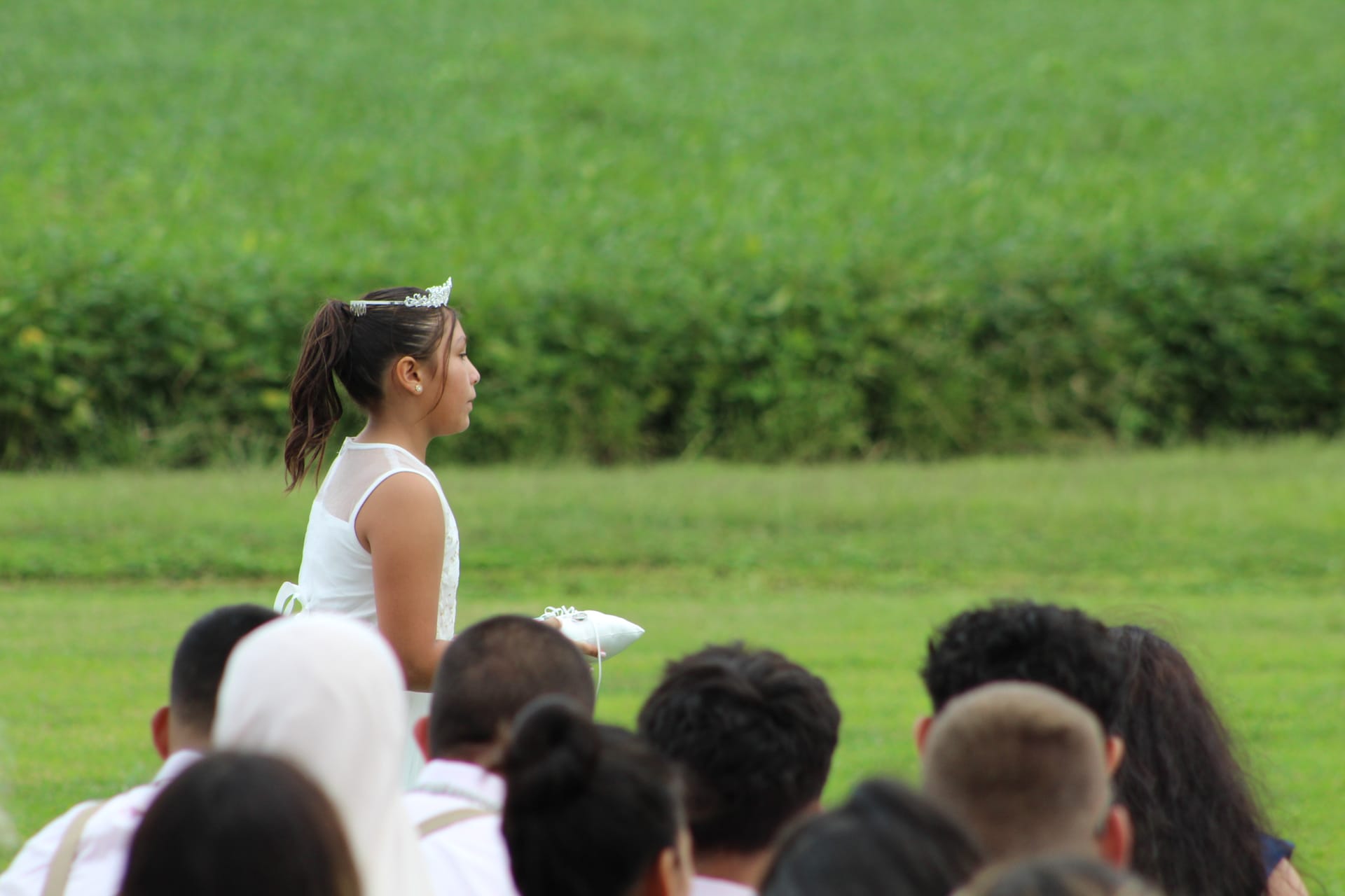 A young flower girl in a white dress and tiara holds a ring pillow during an outdoor wedding ceremony at Scarborough House with green farmland fields in the background.