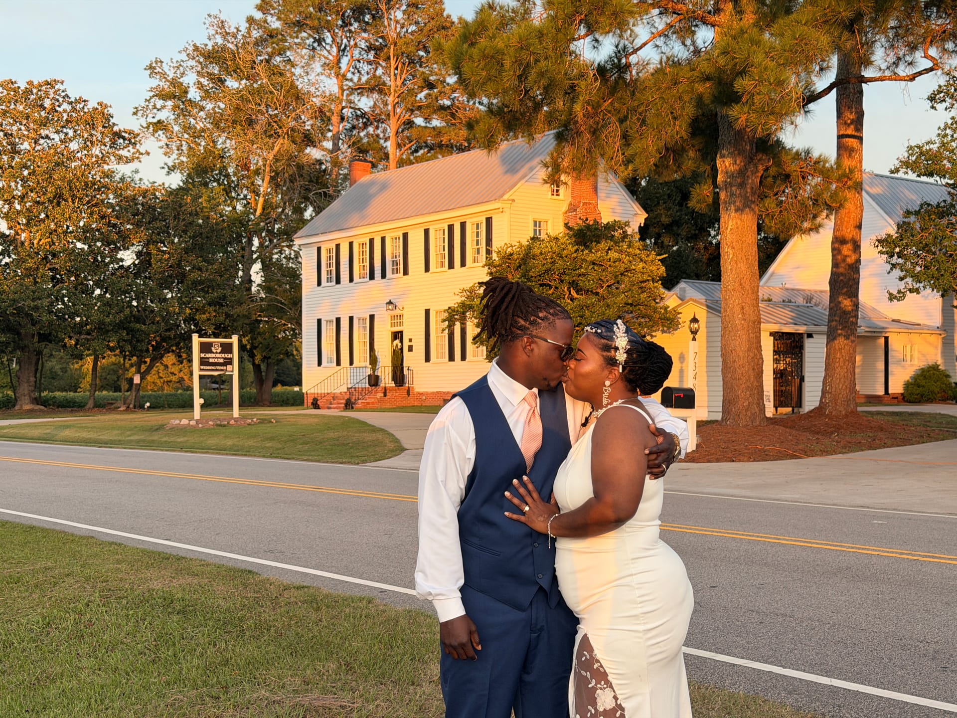 Bride and groom sharing a kiss at golden hour in front of the historic 1821 Scarborough House manor with tall pine trees and warm sunset light.