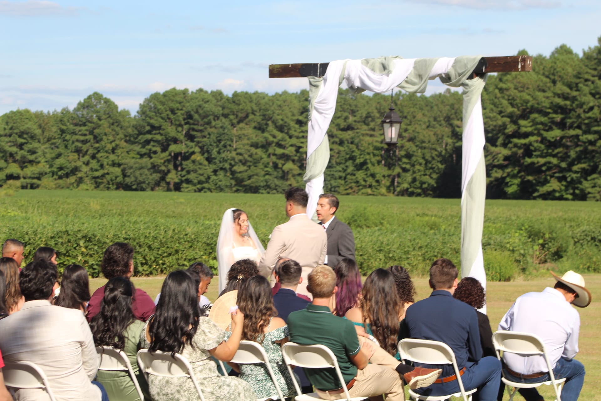 A bride and groom exchange vows beneath a rustic wooden arch draped in white and sage fabric on the ceremony lawn at Scarborough House, surrounded by seated guests with a lush green farmland and treeline in the background.