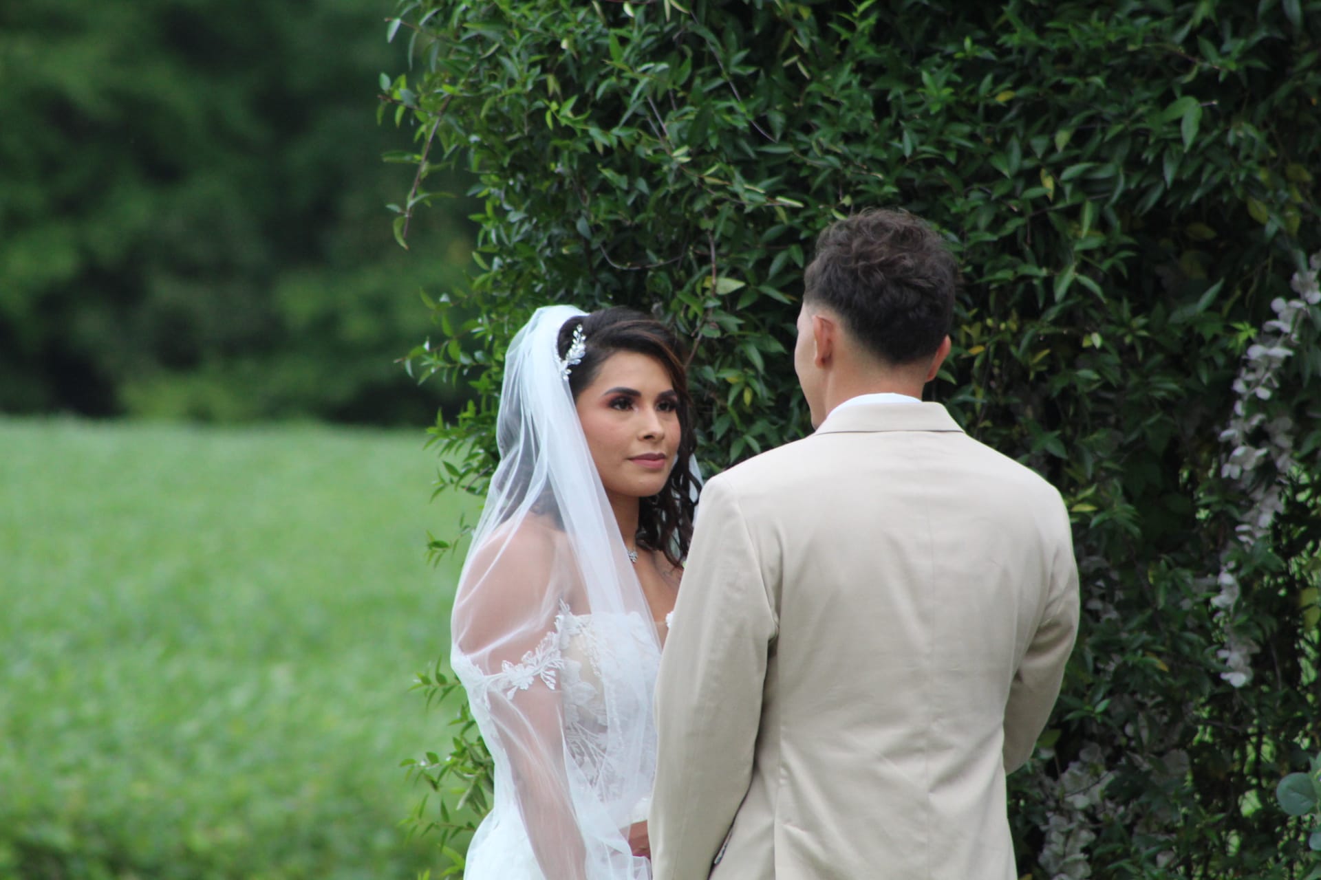 Bride in a lace gown and white veil gazes lovingly at her groom during an outdoor ceremony beside a lush floral and greenery arch at Scarborough House estate.