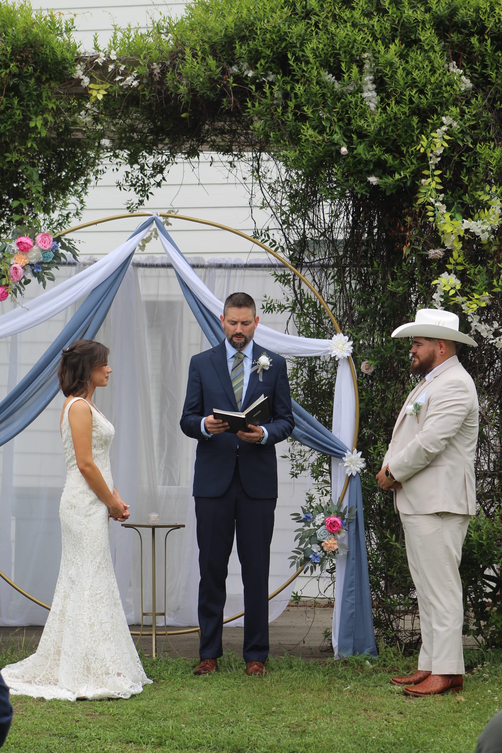 A bride in a white lace gown and groom in a tan suit and white cowboy hat exchange vows before an officiant under a gold circular arch adorned with blue draping and colorful florals on a garden ceremony lawn.