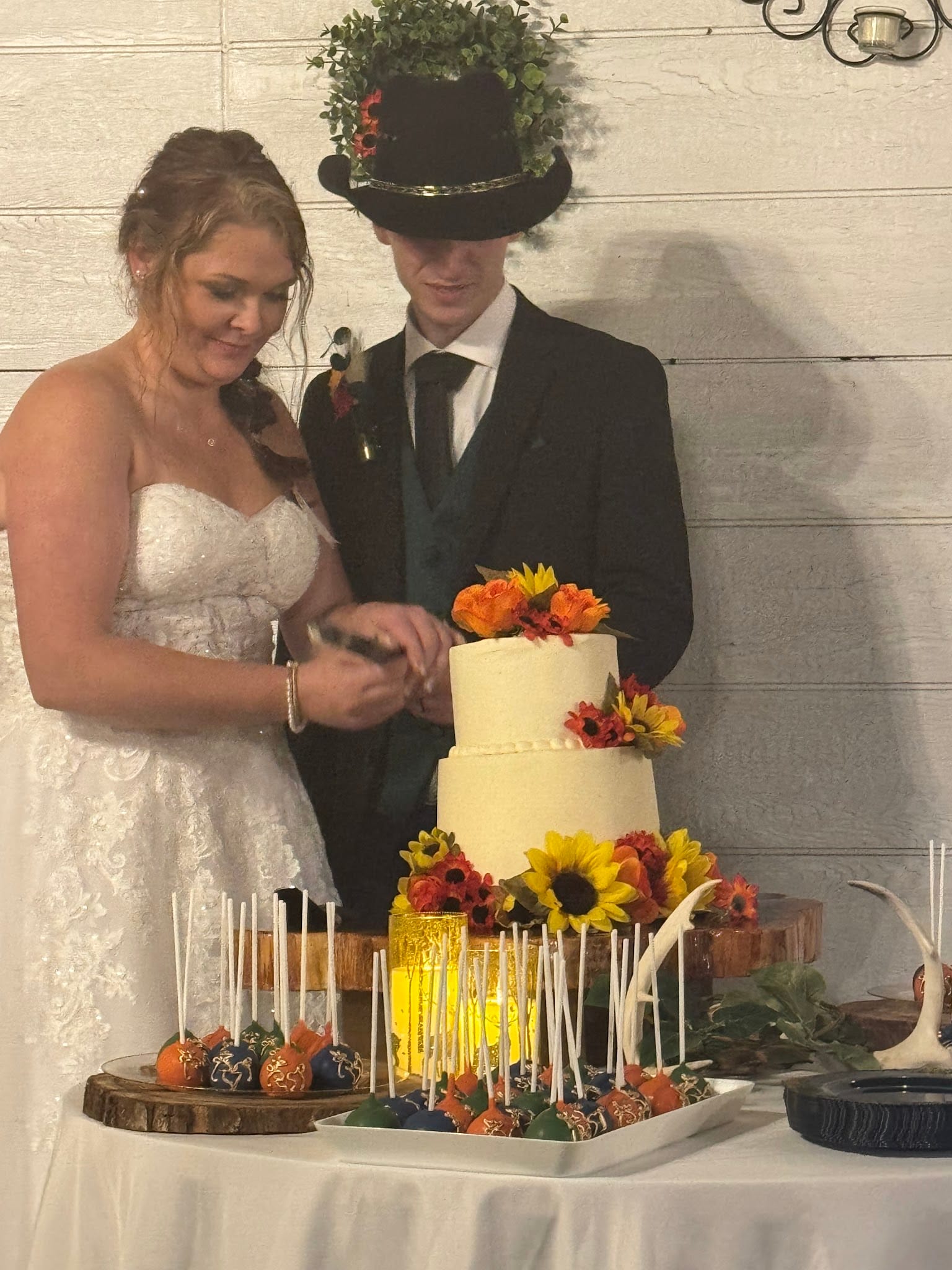 Bride in lace gown and groom in black suit with cowboy hat cut their three-tier white wedding cake adorned with sunflowers and autumn flowers at Scarborough House.