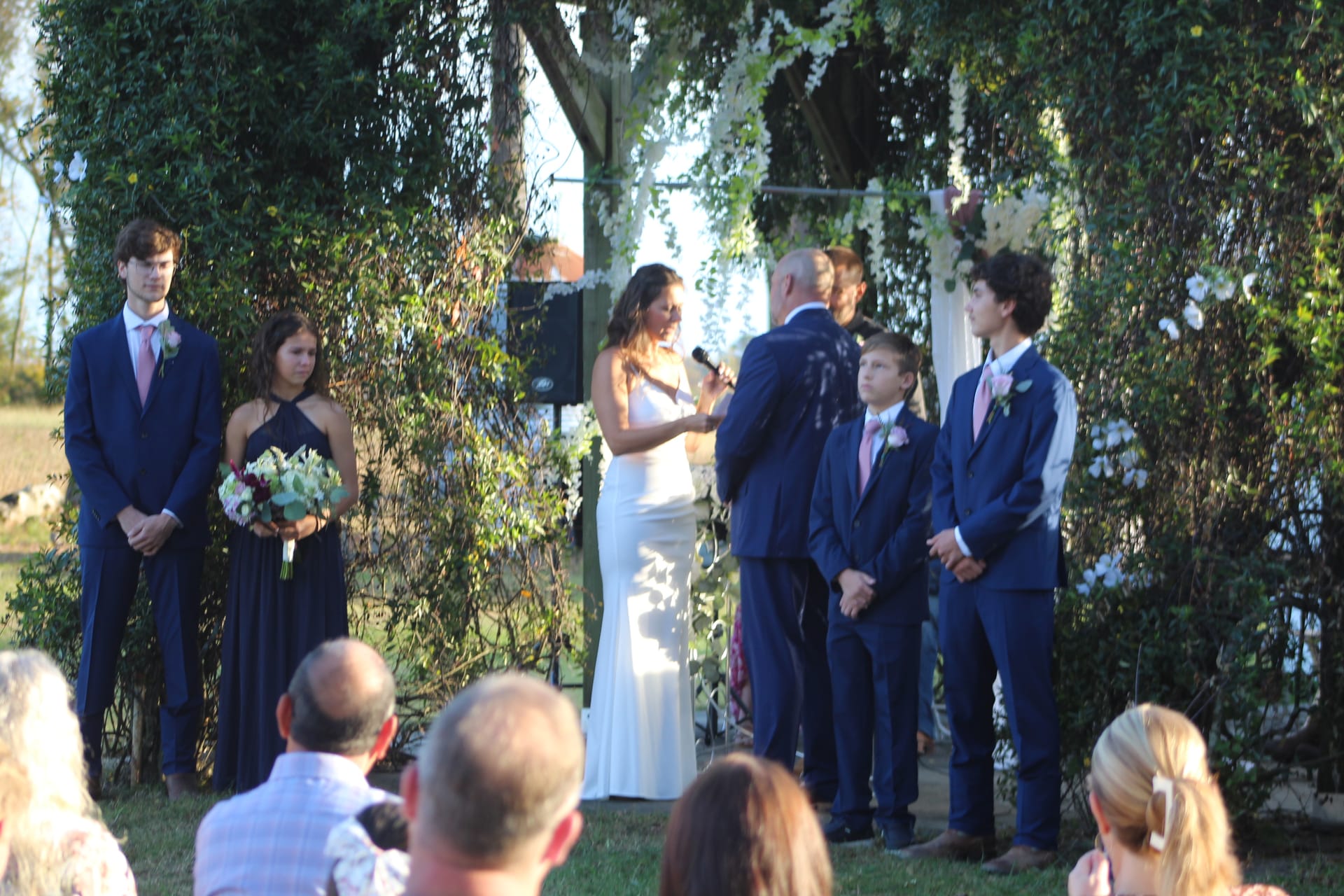 Bride reads vows to groom beneath an ivy-draped archway on the ceremony lawn at Scarborough House, flanked by groomsmen and a bridesmaid holding a floral bouquet.