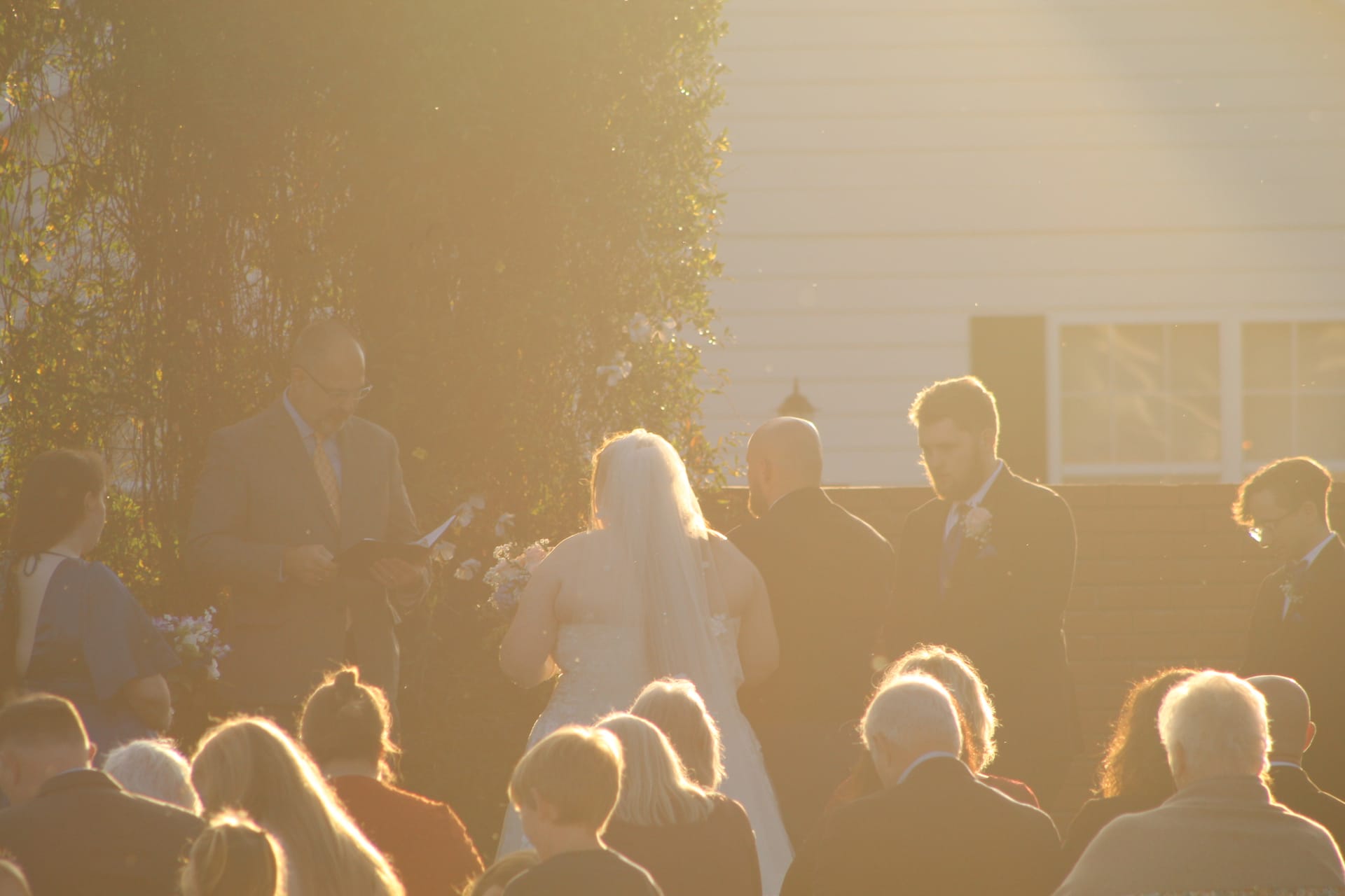 Bride in white gown and veil stands with groom before an officiant during an outdoor golden hour wedding ceremony at Scarborough House historic estate.