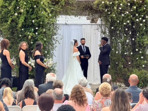 Bride and groom exchange vows beneath a lush floral arch surrounded by bridesmaids in black dresses and seated guests at an outdoor wedding ceremony.