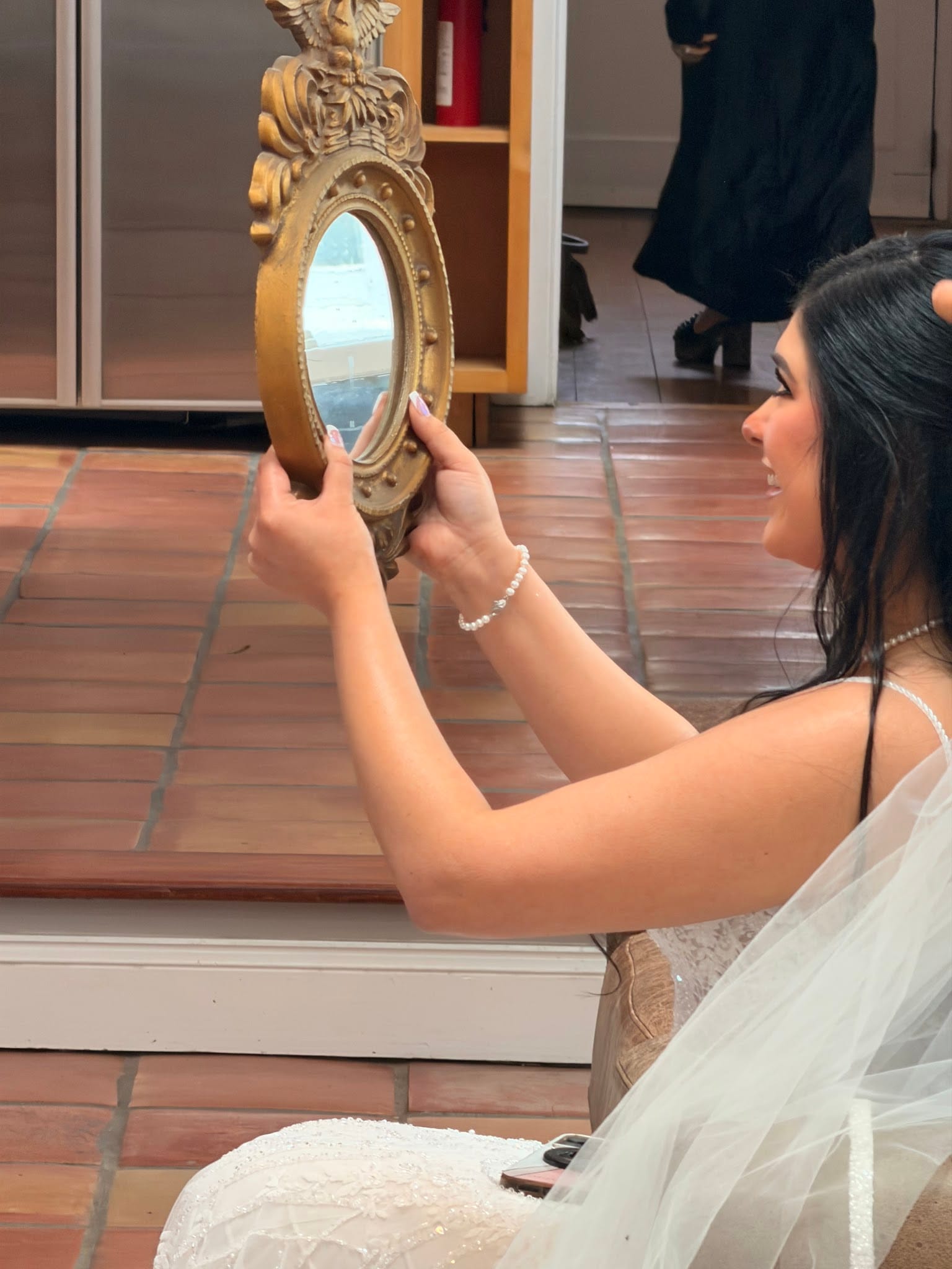 Bride in a lace wedding gown and veil smiles while holding an ornate gold-framed mirror during bridal preparations at Scarborough House.
