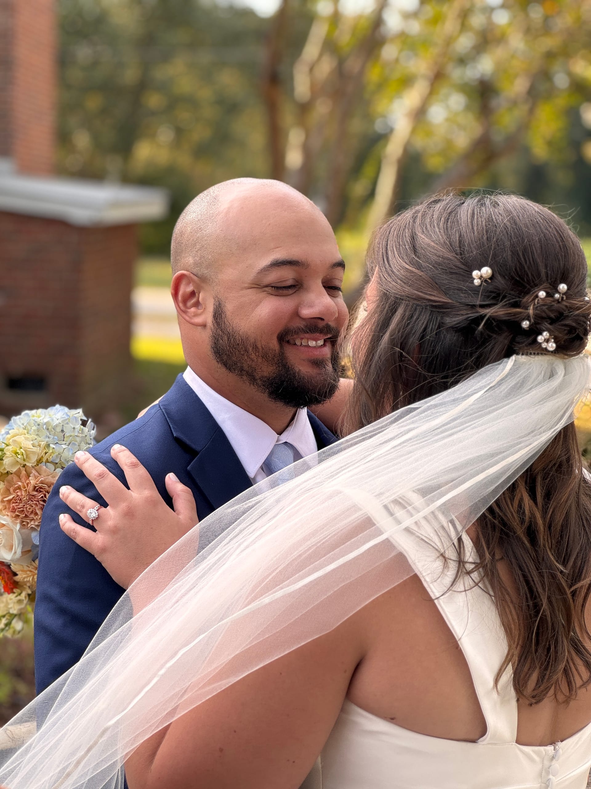 Smiling groom in a navy suit embraces his bride in a white gown and veil adorned with pearl hair pins as she holds a floral bouquet during their outdoor wedding ceremony.
