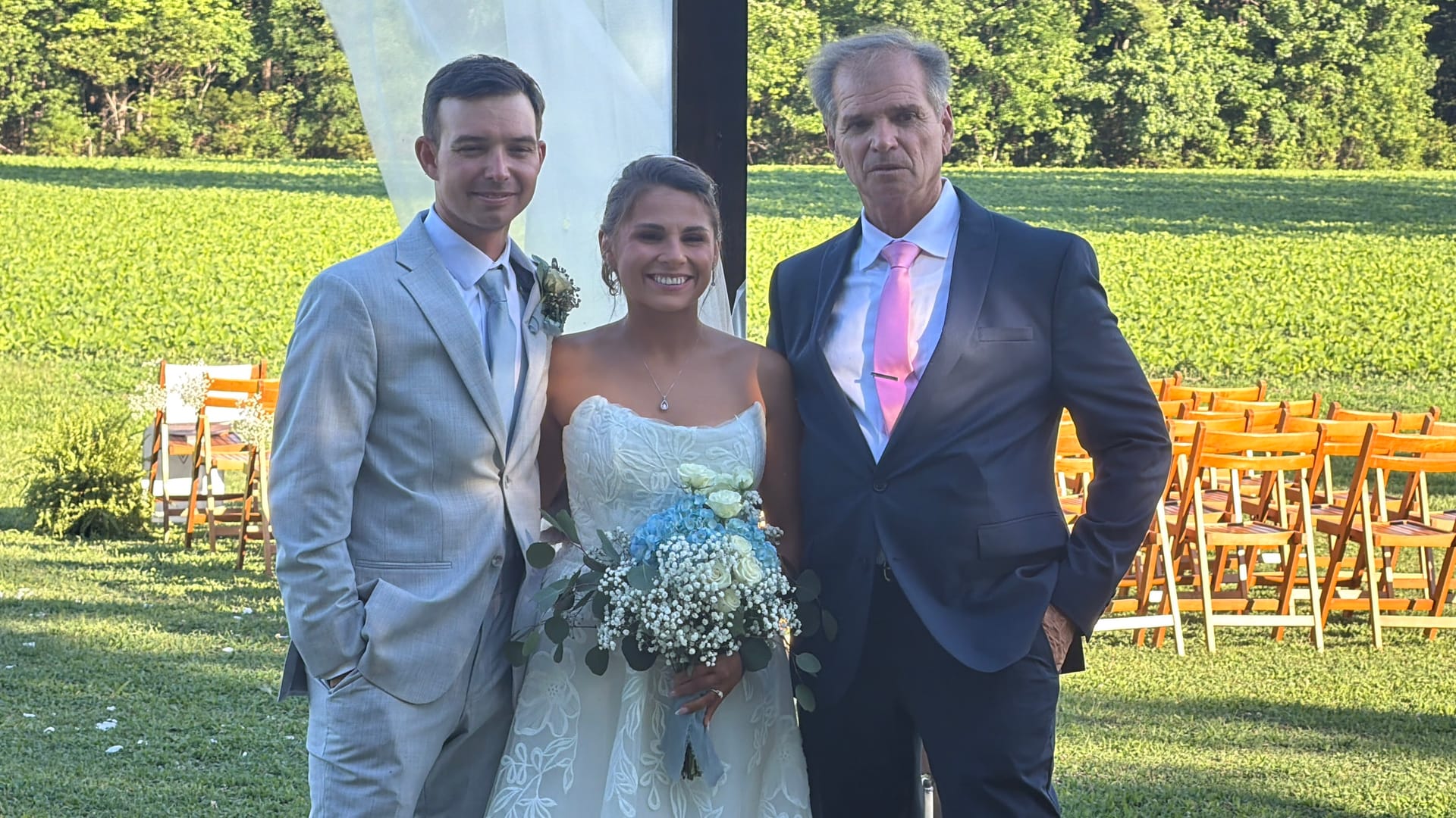 Bride in white gown holding blue and white bouquet poses with groom in light gray suit and an older gentleman in navy suit on the ceremony lawn at Scarborough House with green farmland fields in the background.
