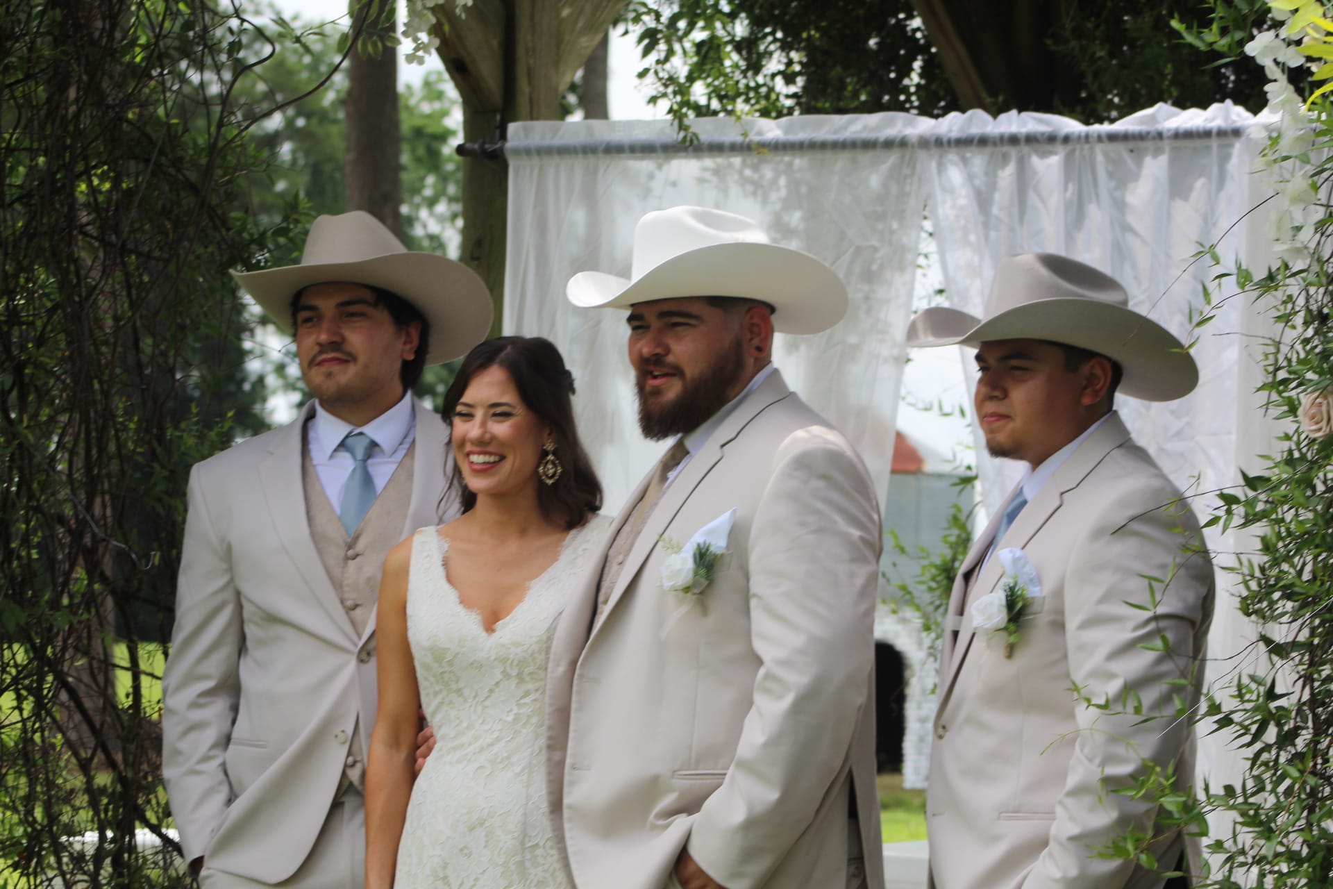 Smiling bride in a lace wedding gown stands with three groomsmen in tan suits and cowboy hats beneath a white draped ceremony arch surrounded by greenery at an outdoor wedding.