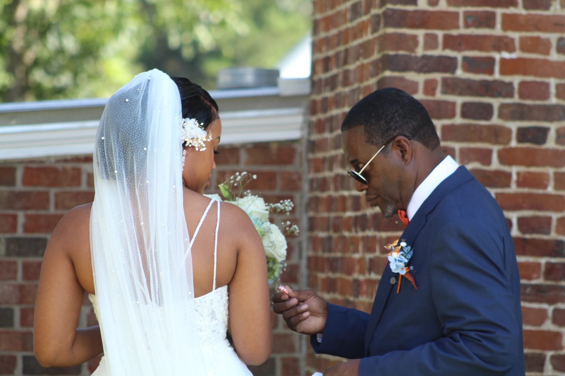 A groom in a navy suit tenderly holds the hands of his bride in a white lace gown and pearl-dotted veil beside a historic brick column at Scarborough House.