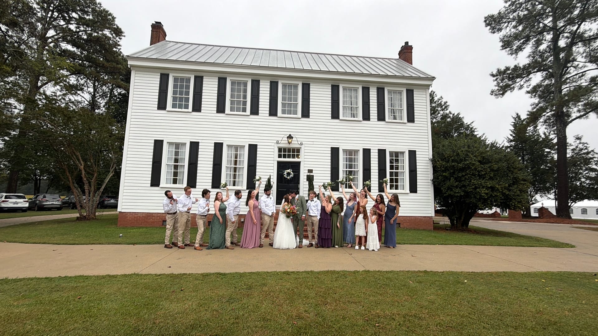 Large wedding party celebrating with arms raised in front of the white 1821 Scarborough House manor on an overcast day.