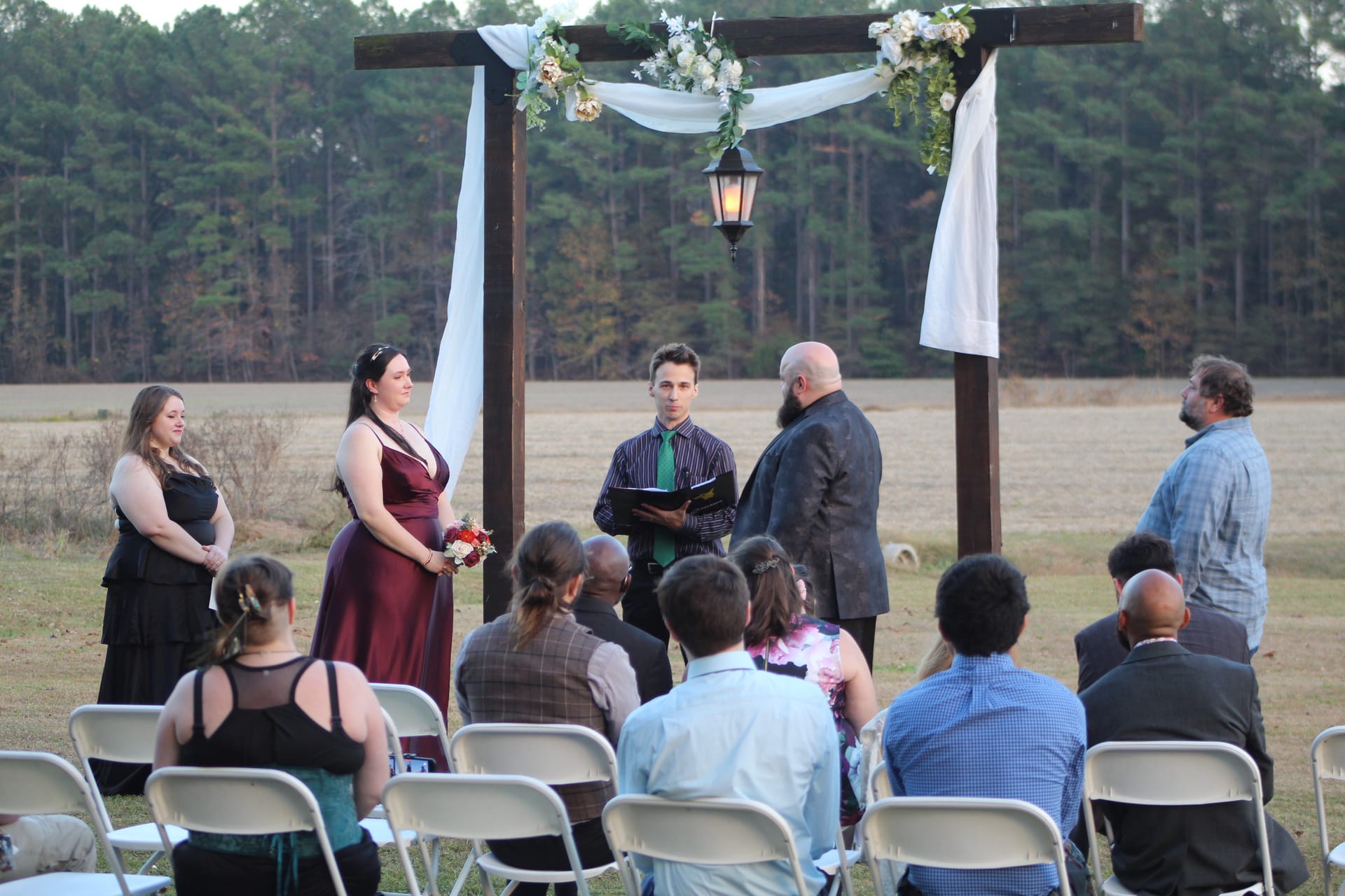 A small outdoor wedding ceremony at dusk beneath a rustic wooden arch adorned with white florals and draped fabric, set against open farmland fields and a pine tree backdrop at Scarborough House.