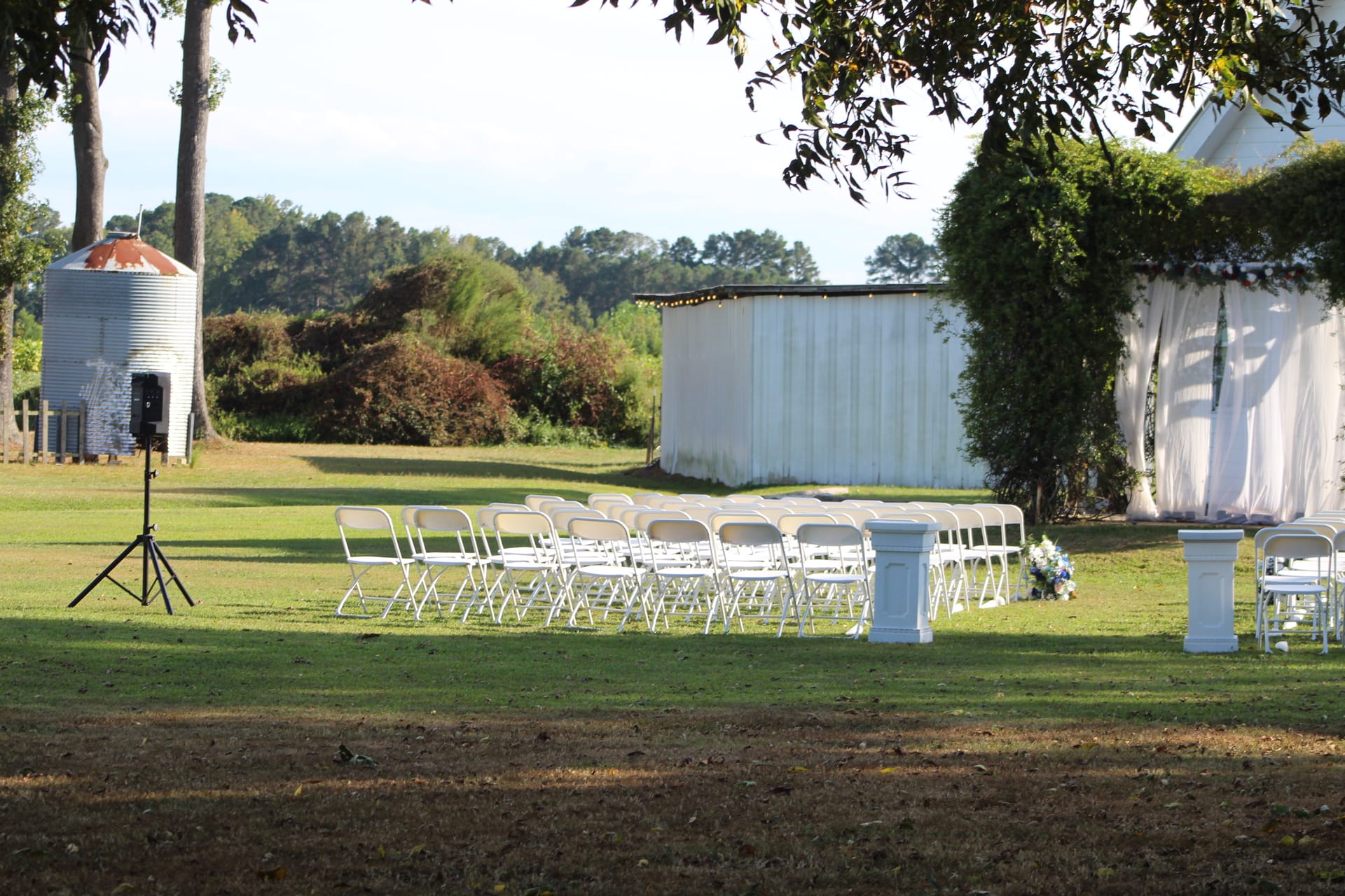 Rows of white folding chairs set up on a sunlit ceremony lawn at Scarborough House with a rustic white barn backdrop, grain silo, and lush green trees in the background.