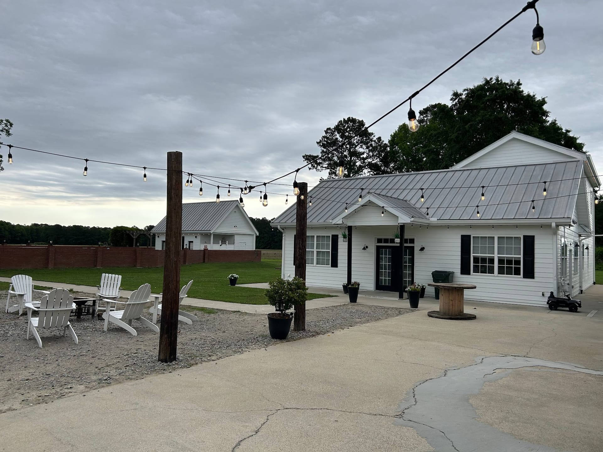 Dusk view of the white farmhouse venue exterior at Scarborough House with glowing string lights strung between wooden posts over a gravel seating area with Adirondack chairs.