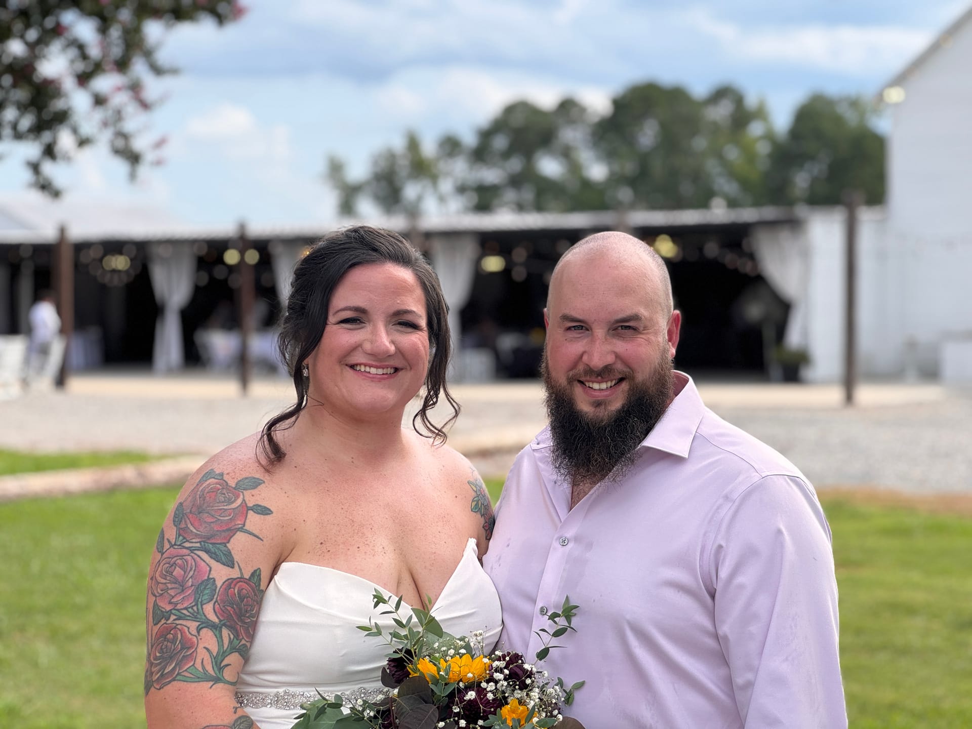 Bride in a strapless white gown holding a sunflower and burgundy bouquet stands smiling beside her bearded groom in a light pink dress shirt on the ceremony lawn at Scarborough House wedding venue.