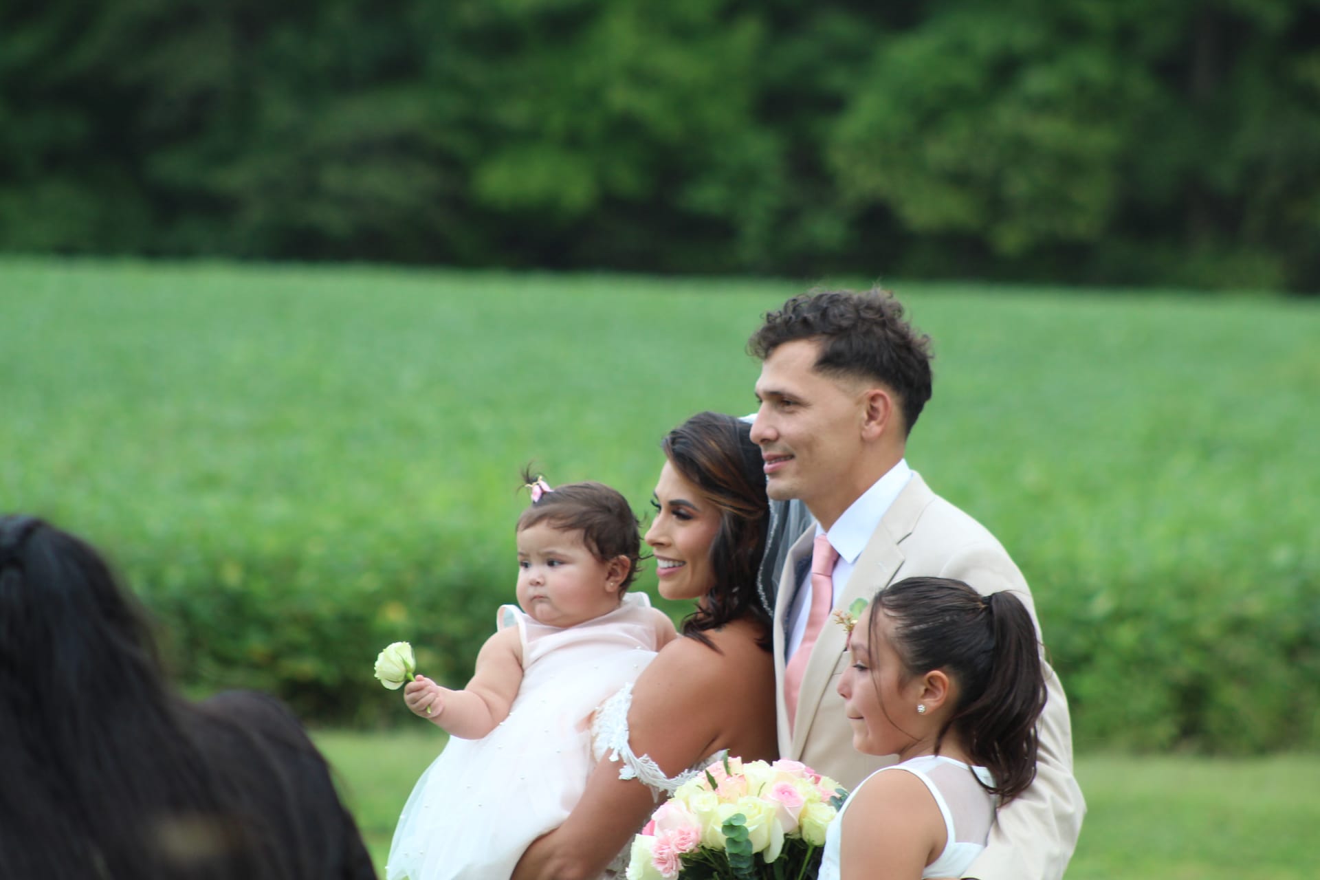 Smiling bride in white gown holds a baby in pink while the groom in a tan suit and pink tie stands beside her with a bridesmaid in front of green farmland fields at Scarborough House.